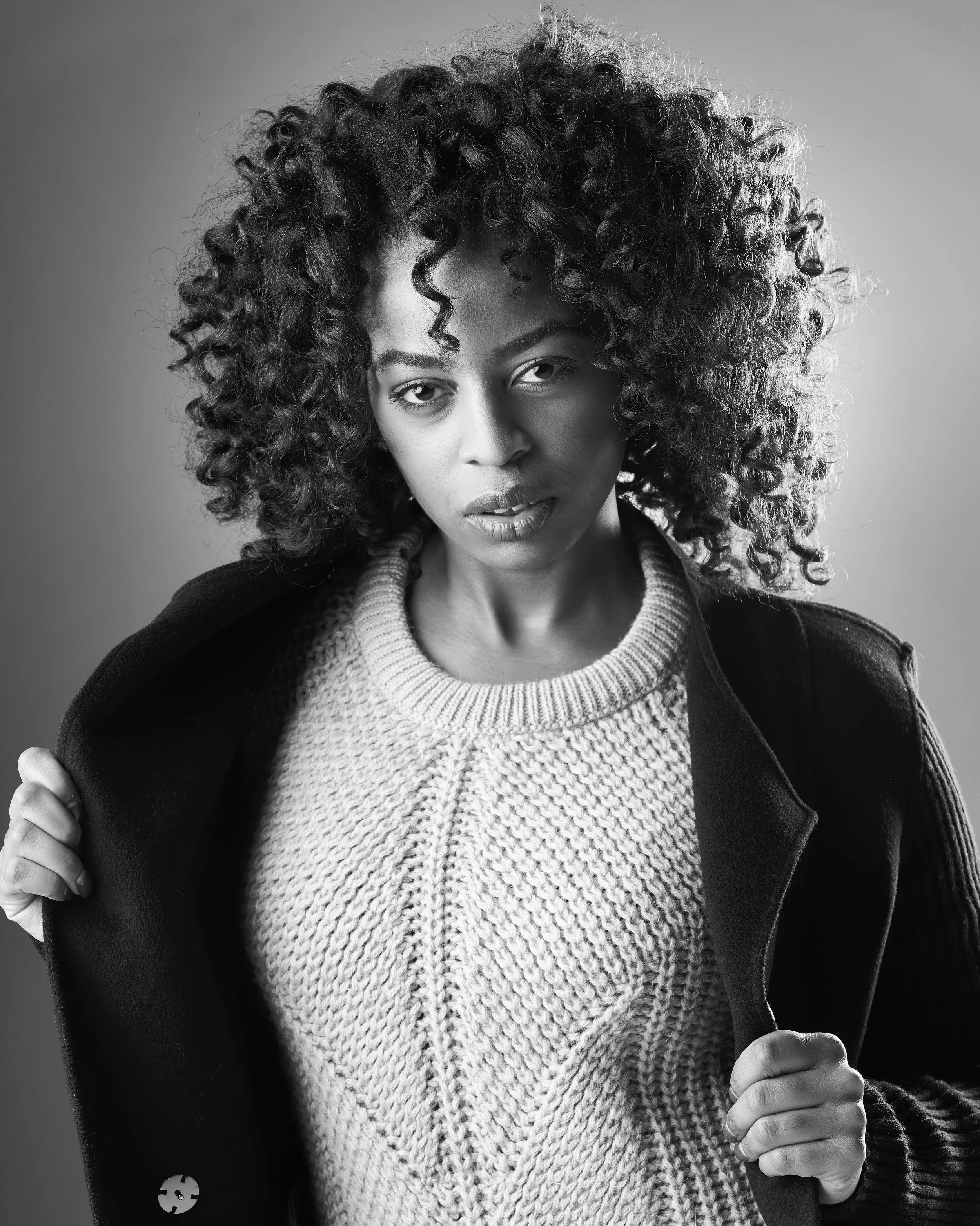 Black and white portrait of a woman with curly hair wearing a sweater and coat, looking directly at the camera.