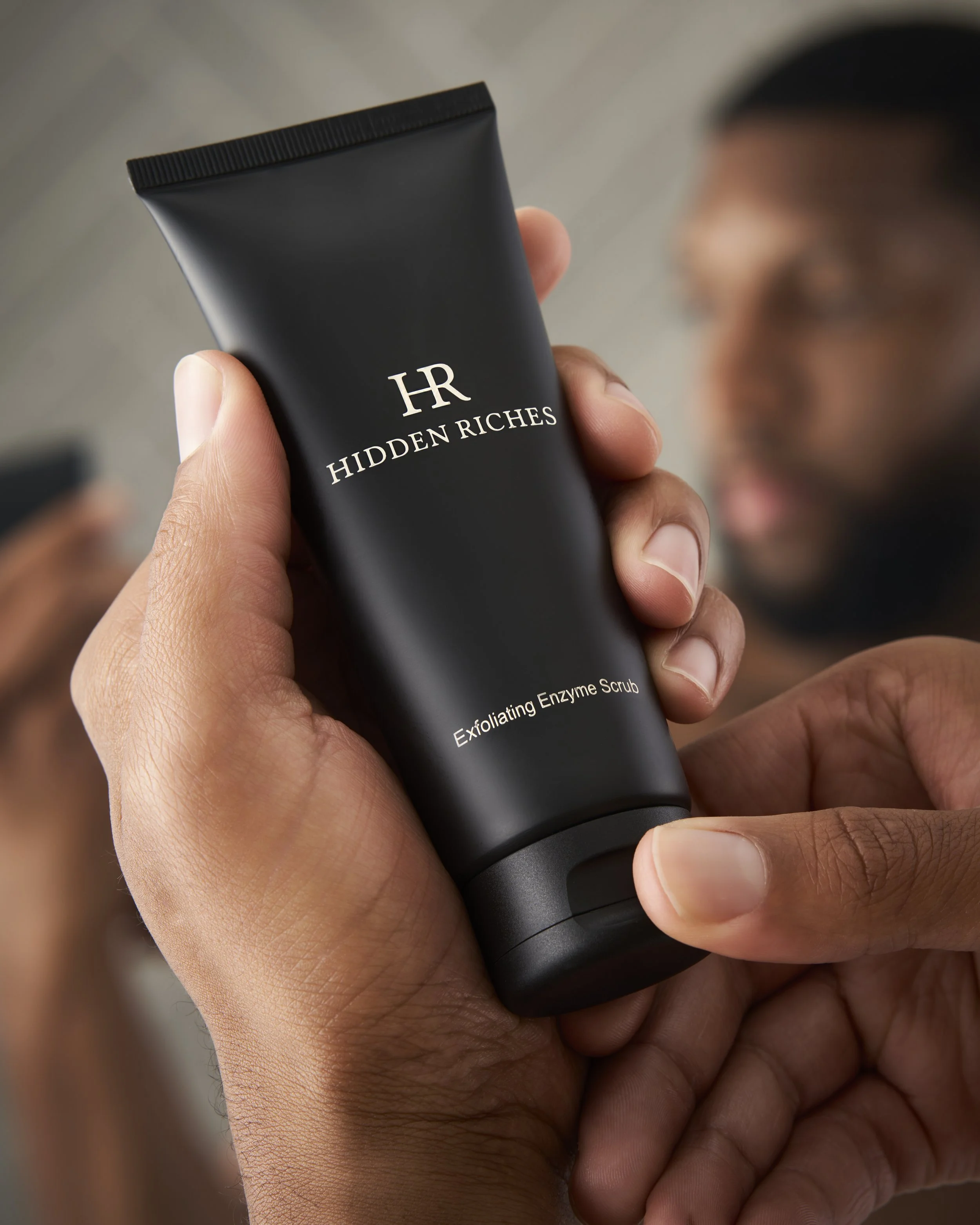 A person holding a black tube of 'Hidden Riches Exfoliating Enzyme Scrub' by HR, with a blurred person in the background.