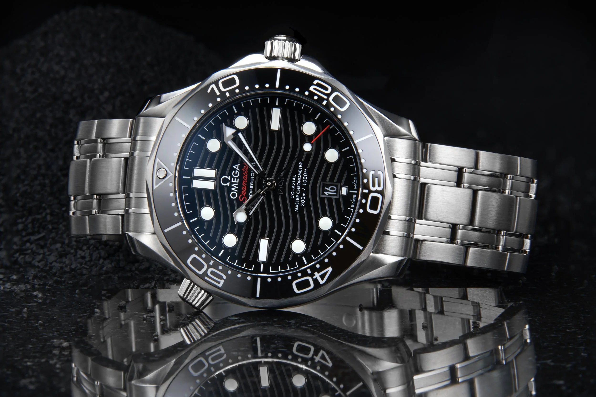 A stainless steel Omega Seamaster Professional dive watch with a black dial, white hour markers, and a rotating bezel with minute markers. The watch is placed on a reflective surface, displaying the time and date.