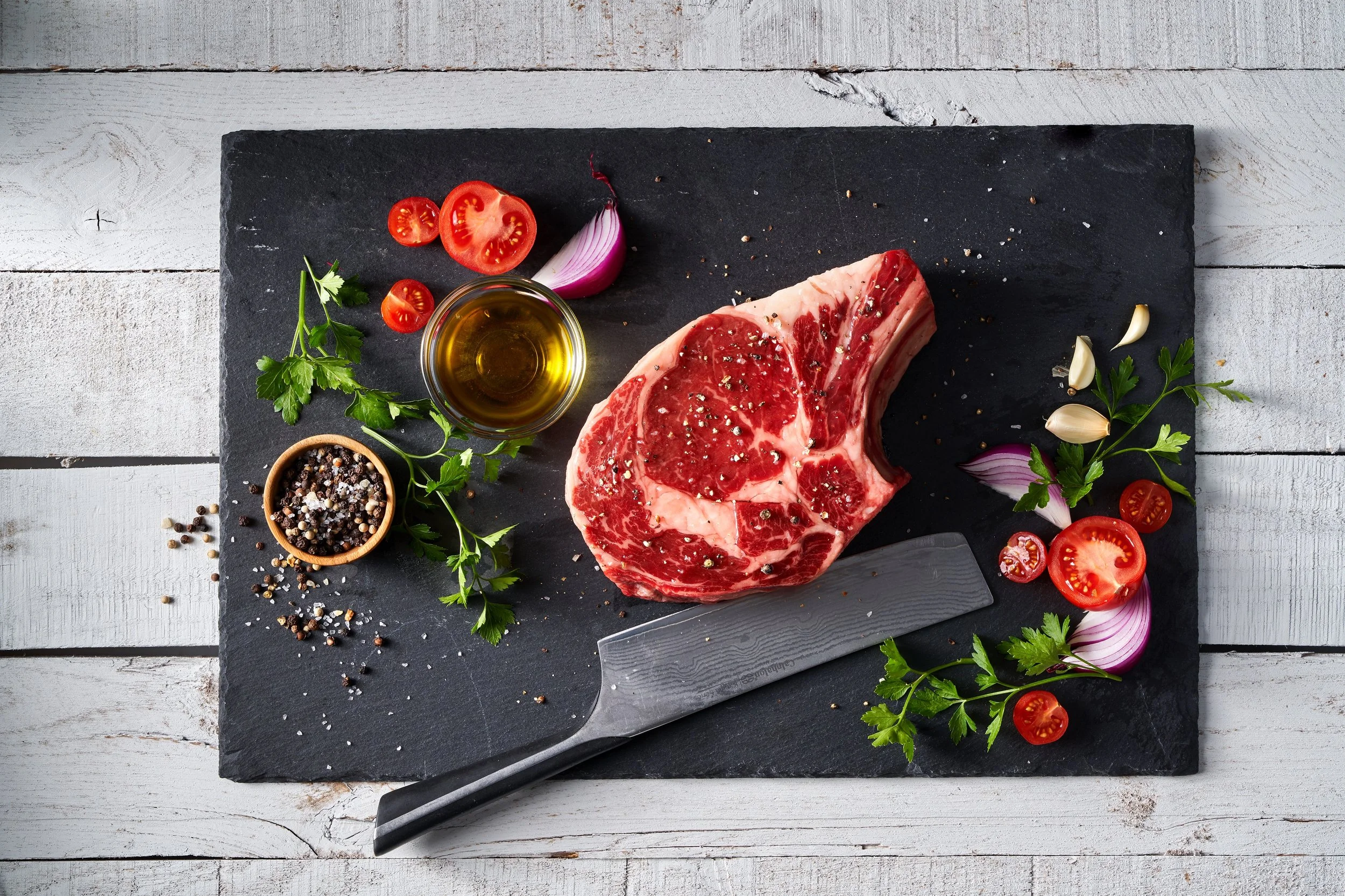 Raw beef ribeye steak seasoned with salt and pepper on a black slate board, surrounded by cherry tomatoes, red onion, garlic cloves, parsley, olive oil, and a jar of mixed peppercorns, with a meat cleaver resting on the board.