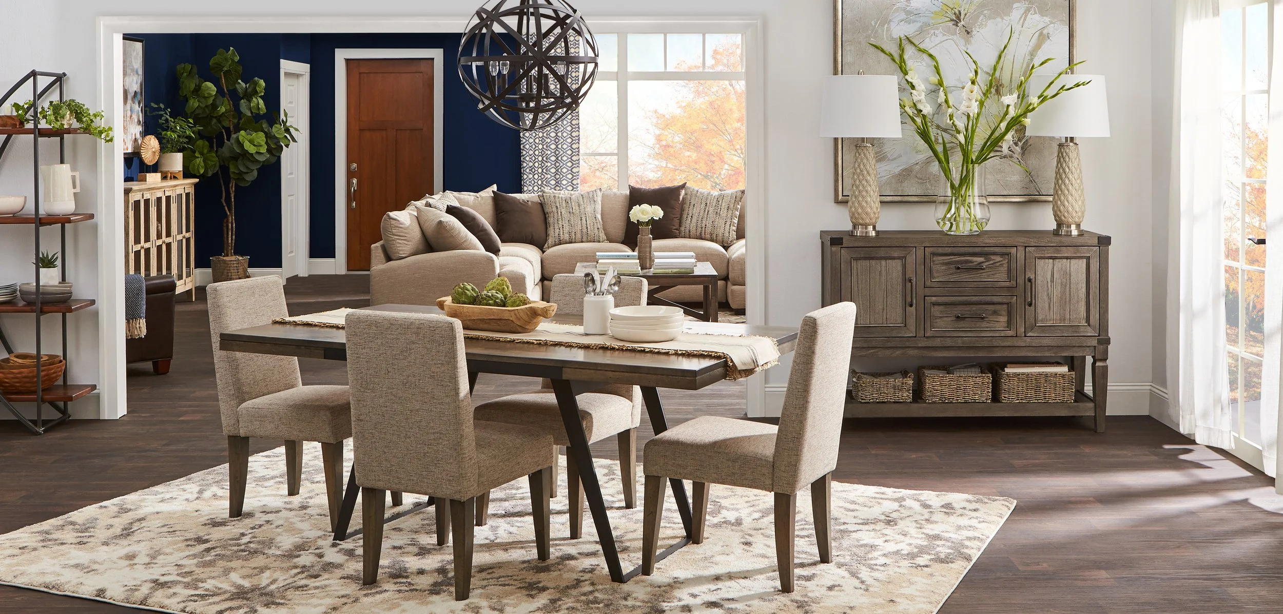 Living and dining room with beige and brown furniture, a dining table with six chairs, a beige sofa, a wooden sideboard with decorative lamps and flowers, open shelving with plants and dishes, dark hardwood floors, large windows with white curtains, 