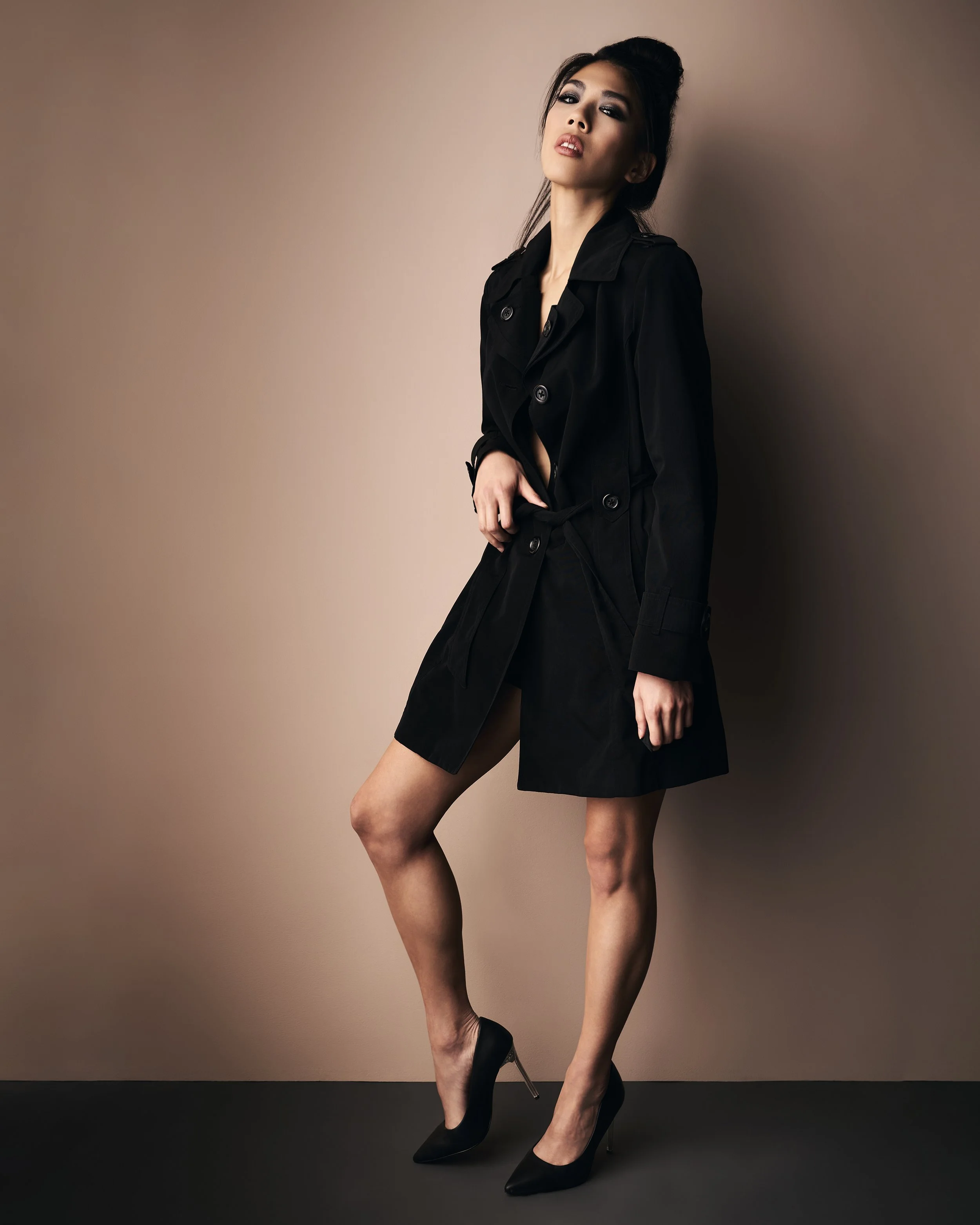 A woman with dark hair styled in an updo, wearing makeup, a black trench coat and black high heels, standing against a neutral background.