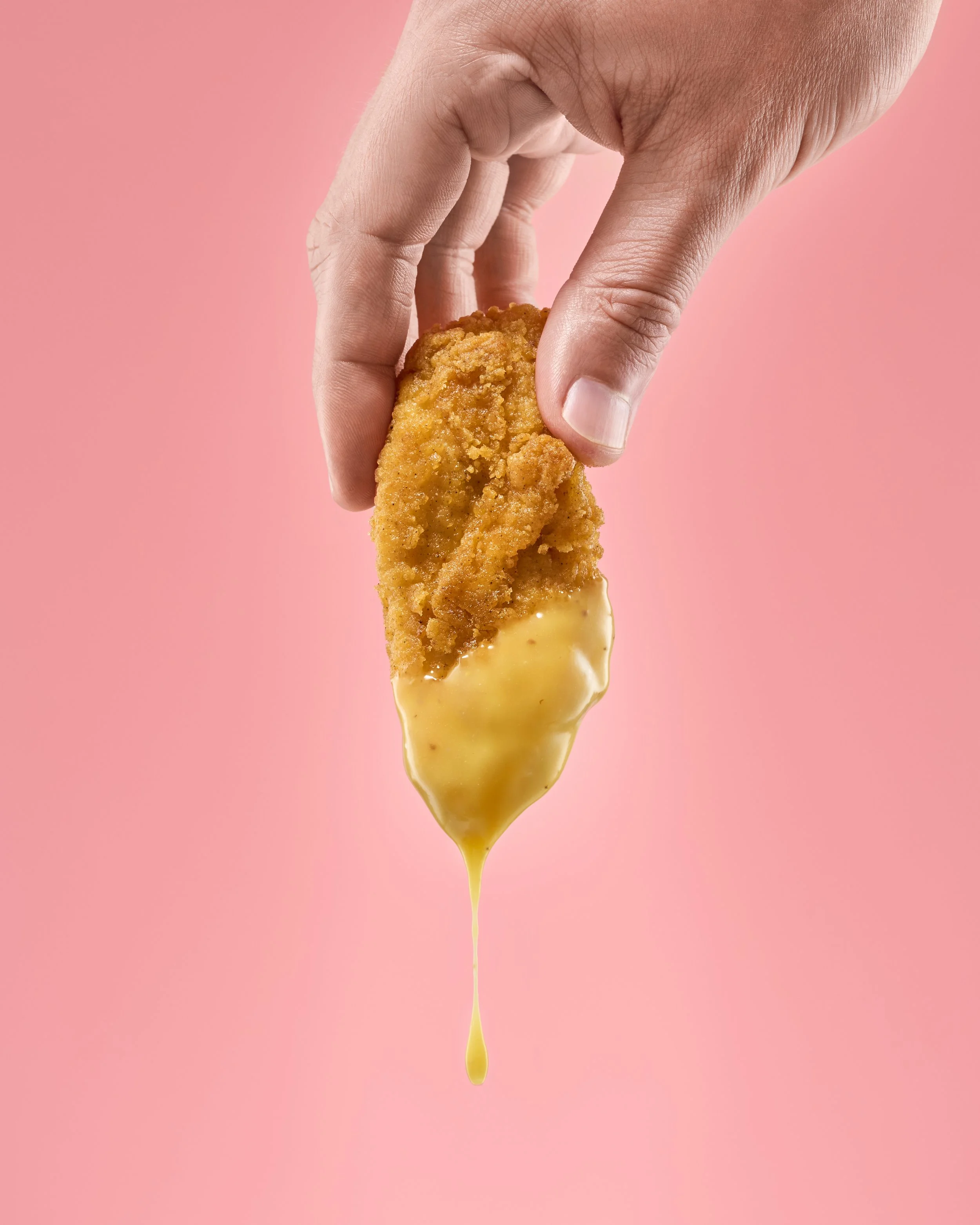 Hand holding a fried chicken tender with melted cheese dripping from it against a pink background.