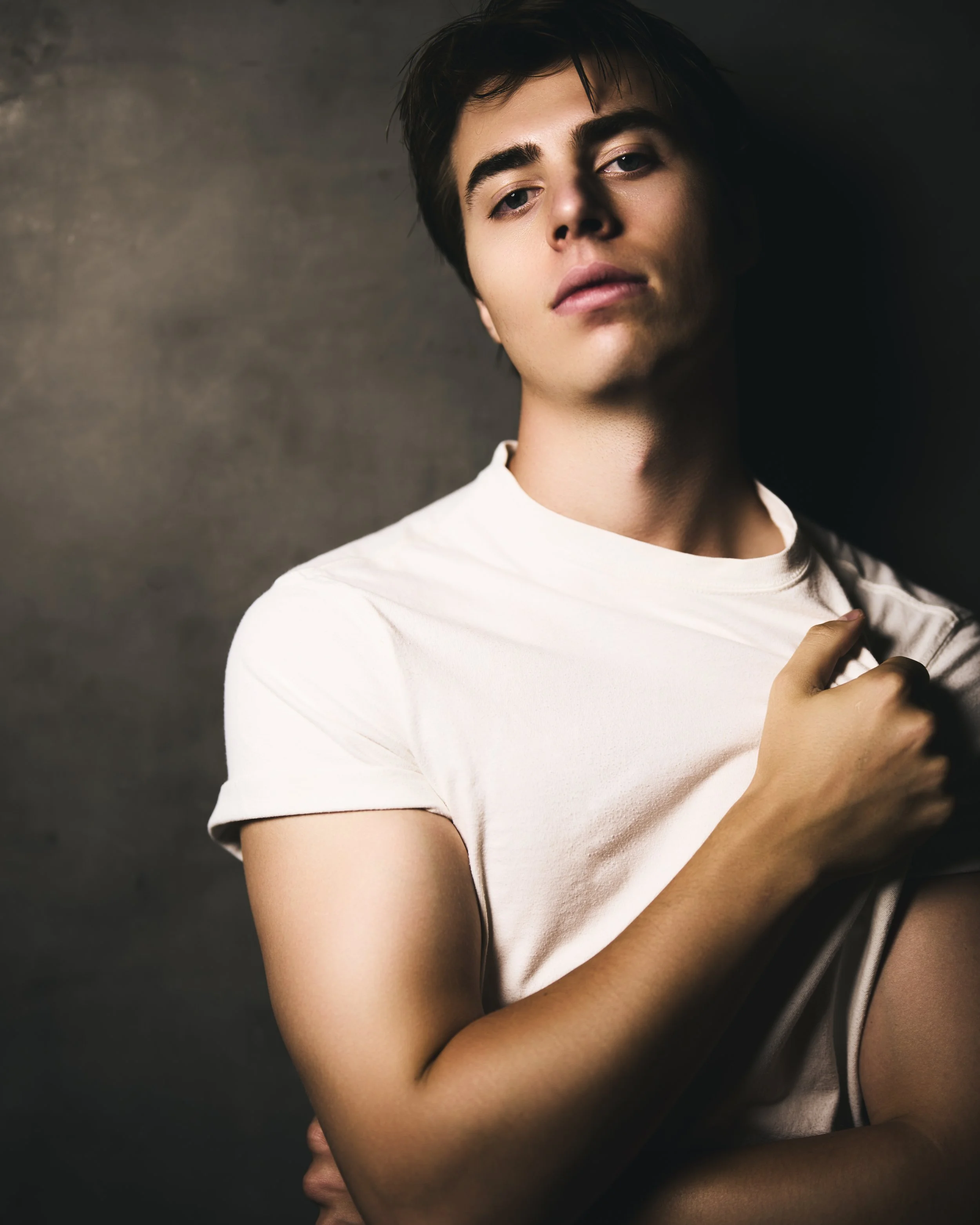 Close-up of a young man with dark hair and fair skin, wearing a white shirt, looking at the camera with a serious expression, against a dark background.