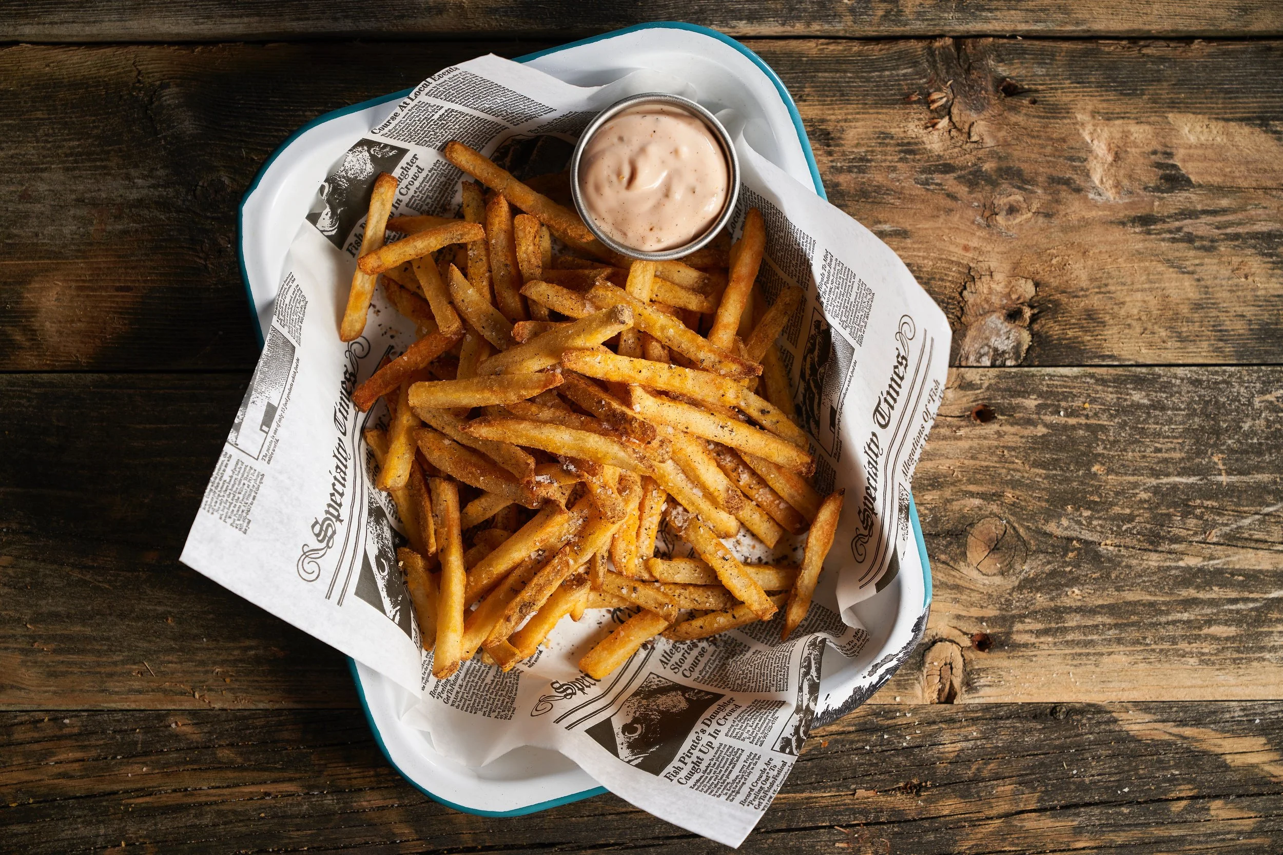Basket of French fries with a small cup of creamy dipping sauce, served on newspaper-lined tray on a rustic wooden table.