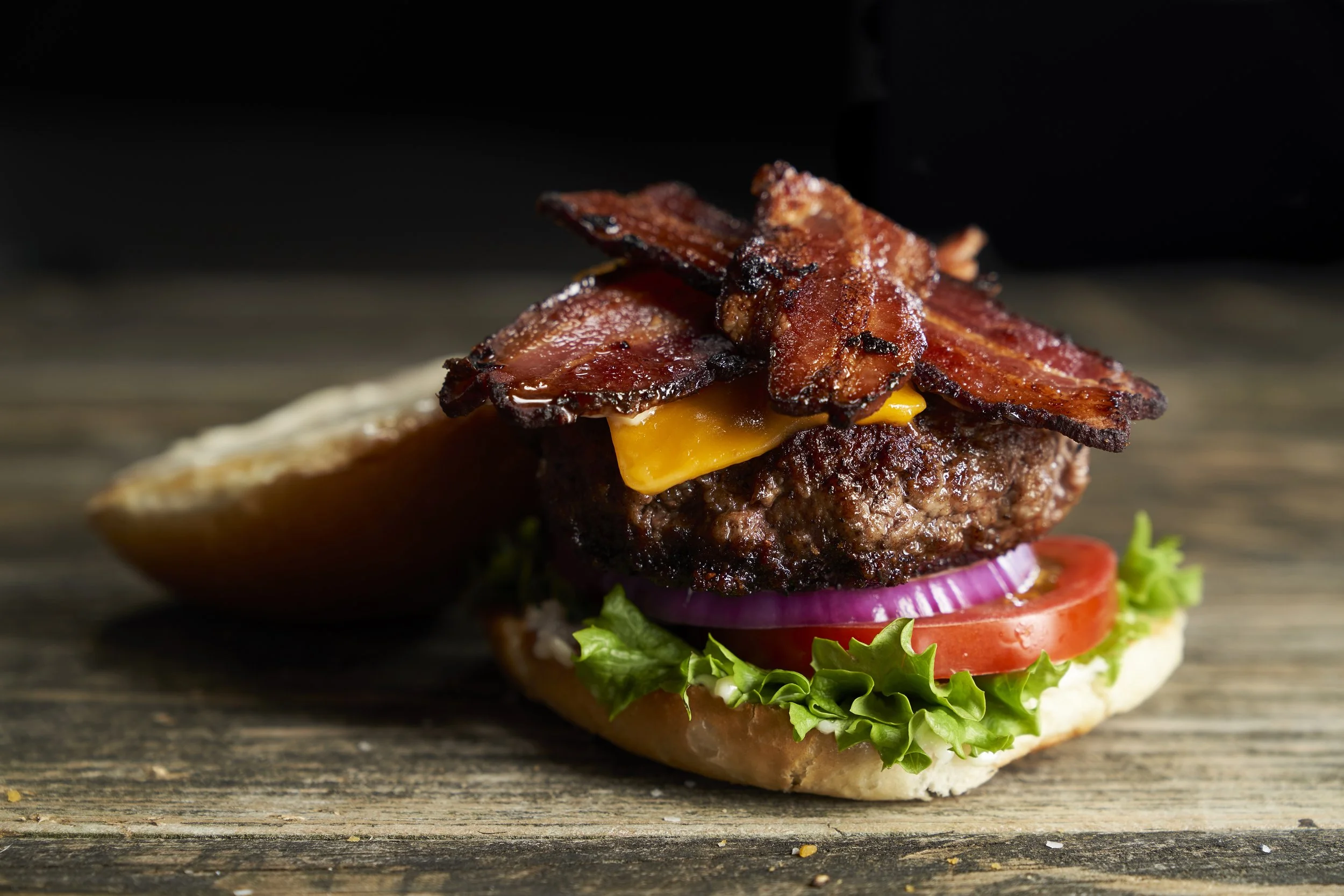 Close-up of a bacon cheeseburger with lettuce, tomato, onion, cheddar cheese, a beef patty, and crispy bacon on a bun, on a wooden surface.