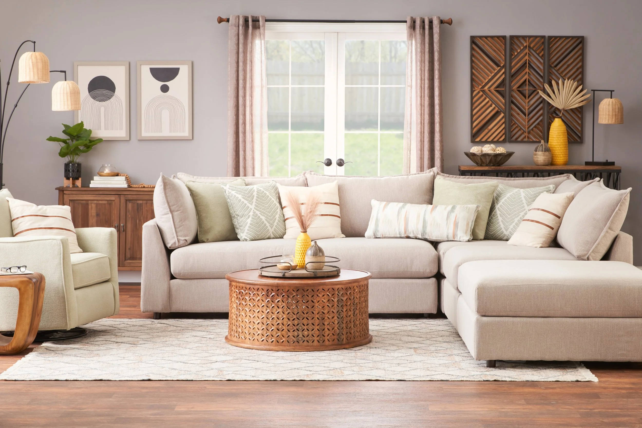 Living room with beige sectional sofa, green and patterned throw pillows, round wooden coffee table with decorative vases, beige armchair, wooden sideboard, abstract wall art, window with pink curtains, and decorative items on shelves.