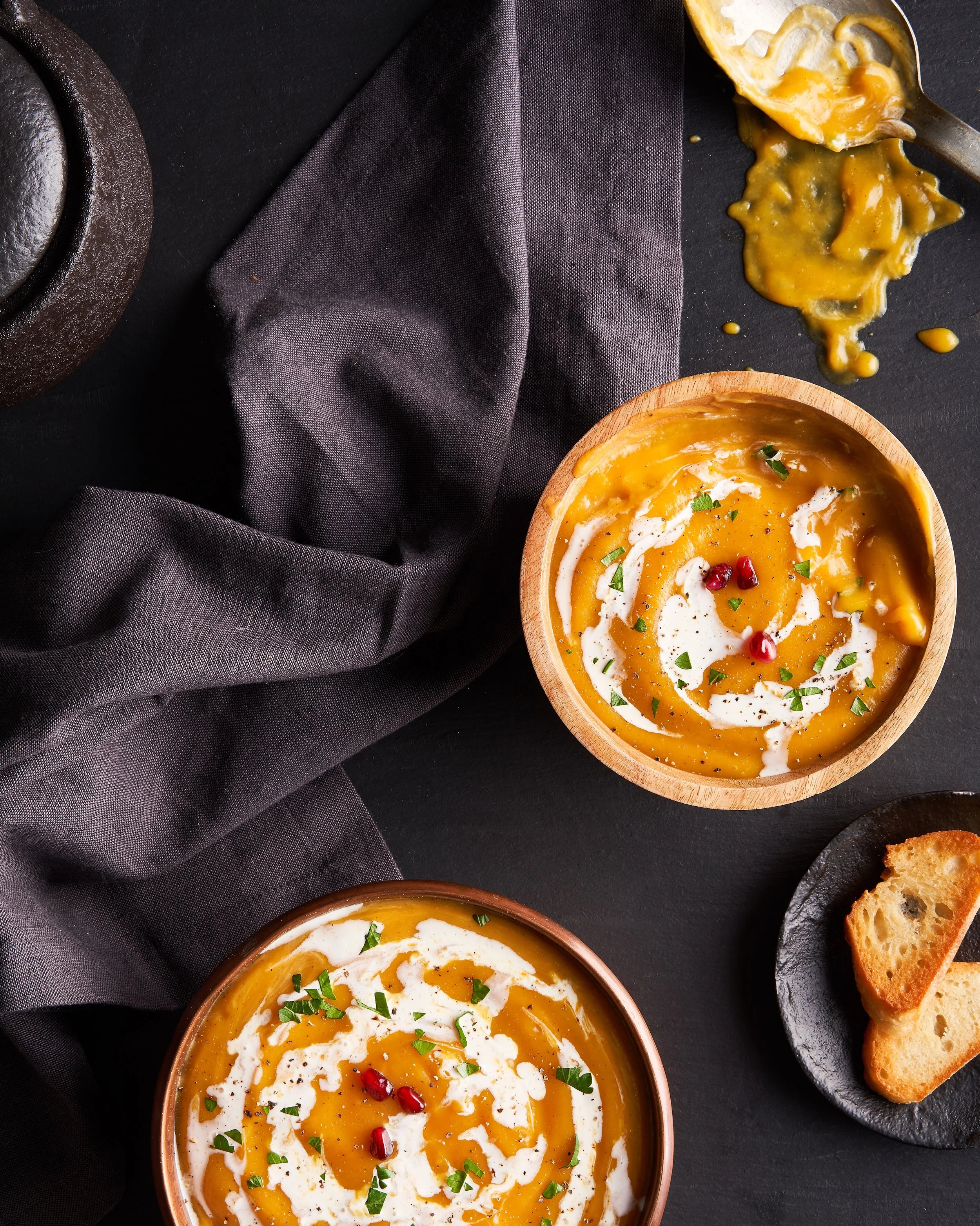 Two bowls of creamy butternut squash soup garnished with cream, pomegranate seeds, and herbs, with slices of bread on a black plate, a spoon with soup and gravy spill, black napkin, and a cast iron pot in the background.