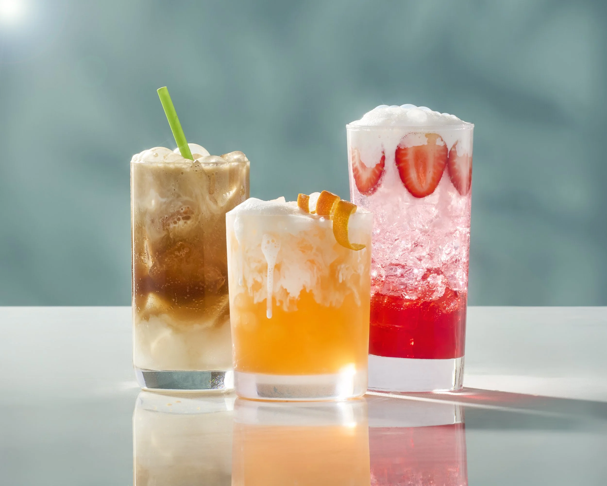 Three colorful cocktails with ice and fruit garnishes on a reflective surface against a blurred light blue background.