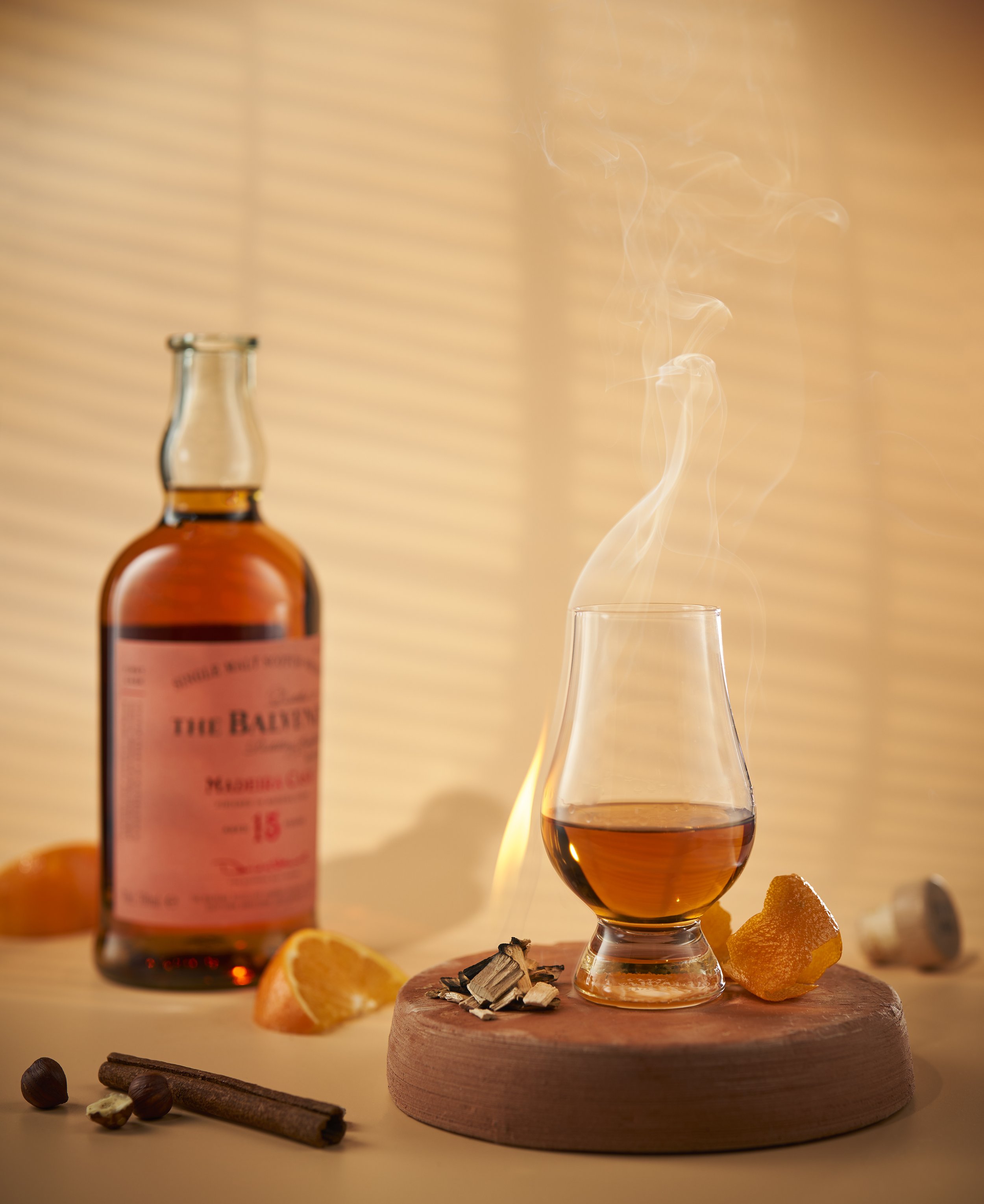 A glass of whiskey on a wooden coaster with orange slices, cinnamon stick, nuts, and other spices. A bottle of whiskey in the background, with warm lighting and a blurred background.