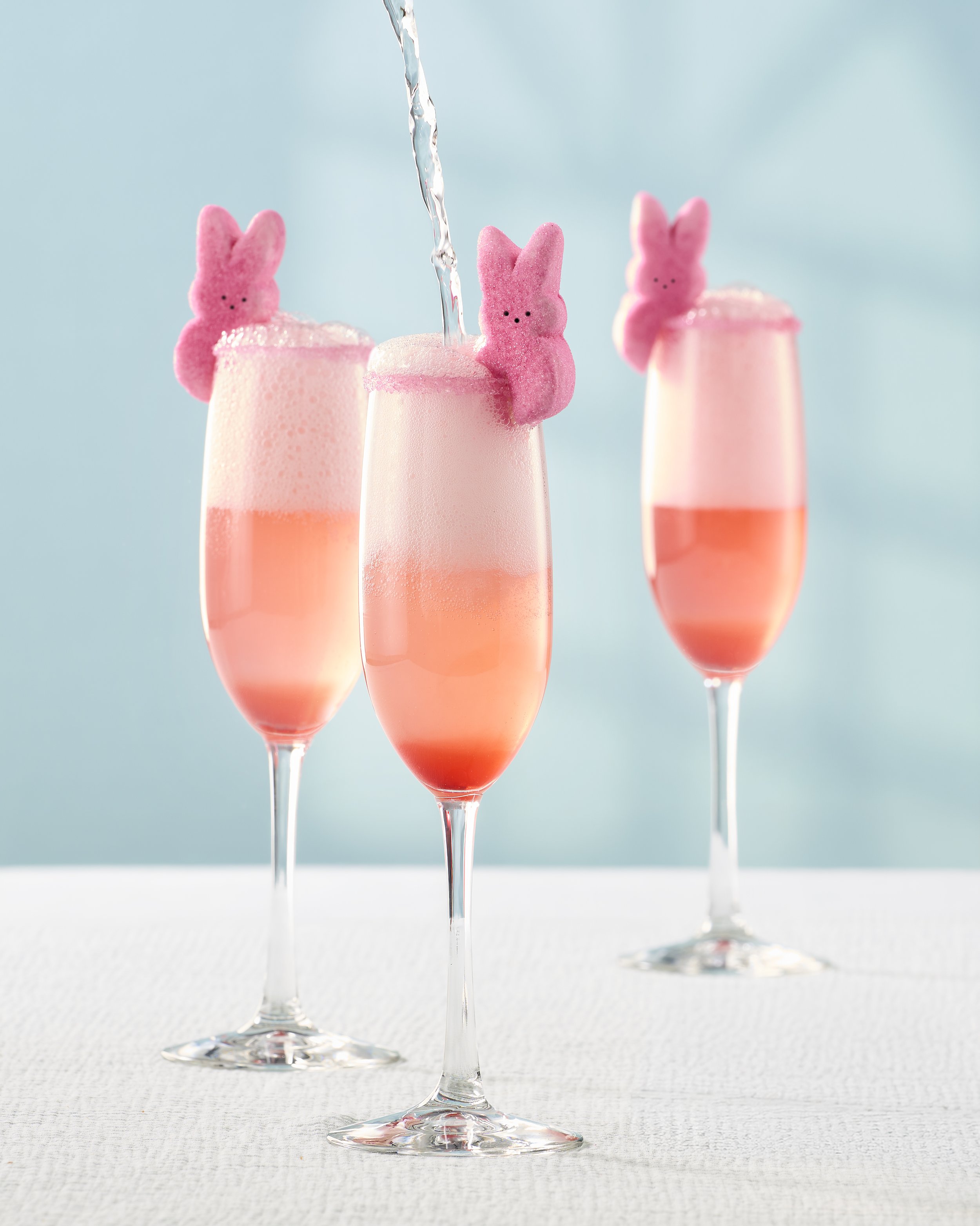 Three champagne flutes filled with pink bubbly drink, each decorated with a pink bunny-shaped marshmallow on the rim, with one glass being filled with water.