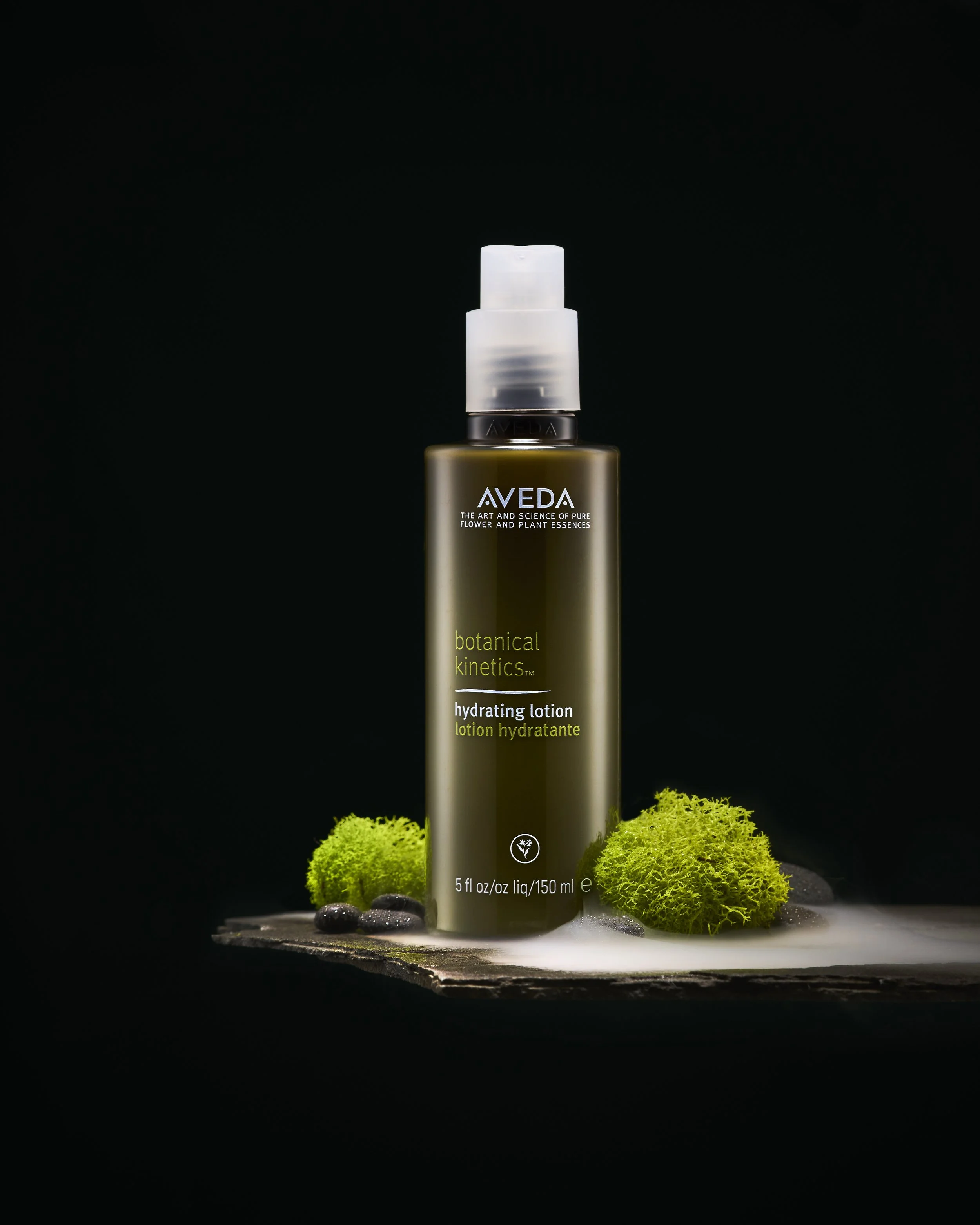 A bottle of Aveda botanical kinetics hydrating lotion with green moss and black stones on a dark background.