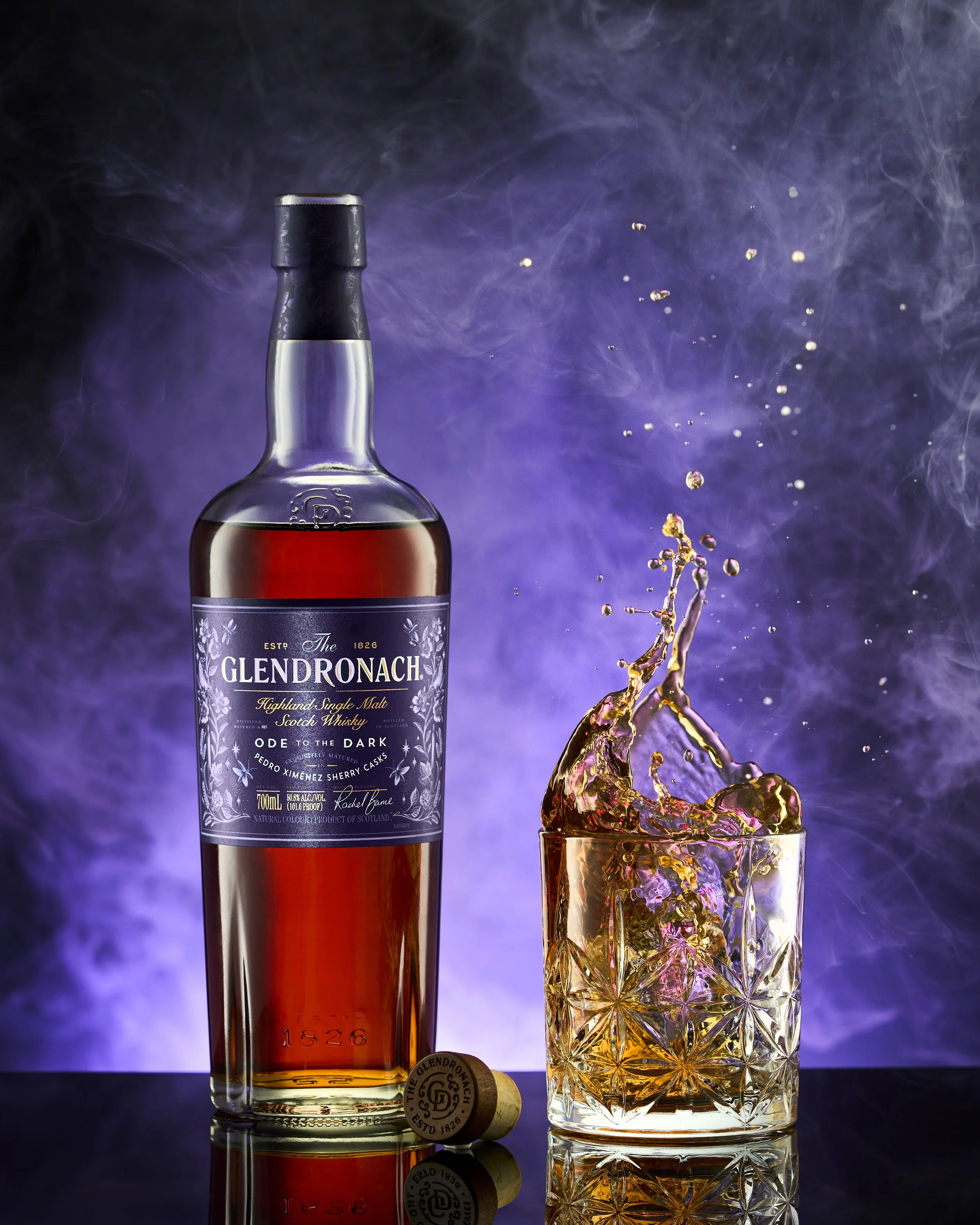 A bottle of Glendronach Highland Single Malt Scotch Whisky next to a glass of whiskey with ice and a splash captured mid-air, alongside a cork with the Glendronach logo, on a reflective surface with a purple smoke background.