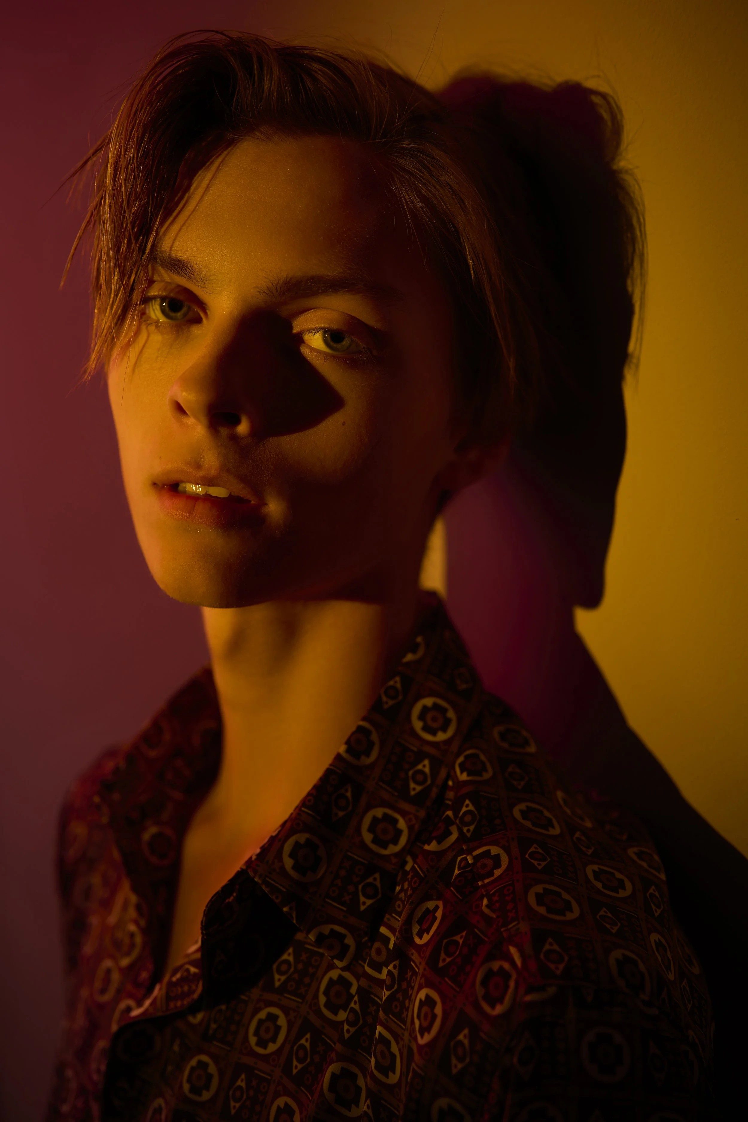 Close-up of a young man with short, tousled hair, wearing a patterned shirt, lit by colorful lighting creating shadows on his face.