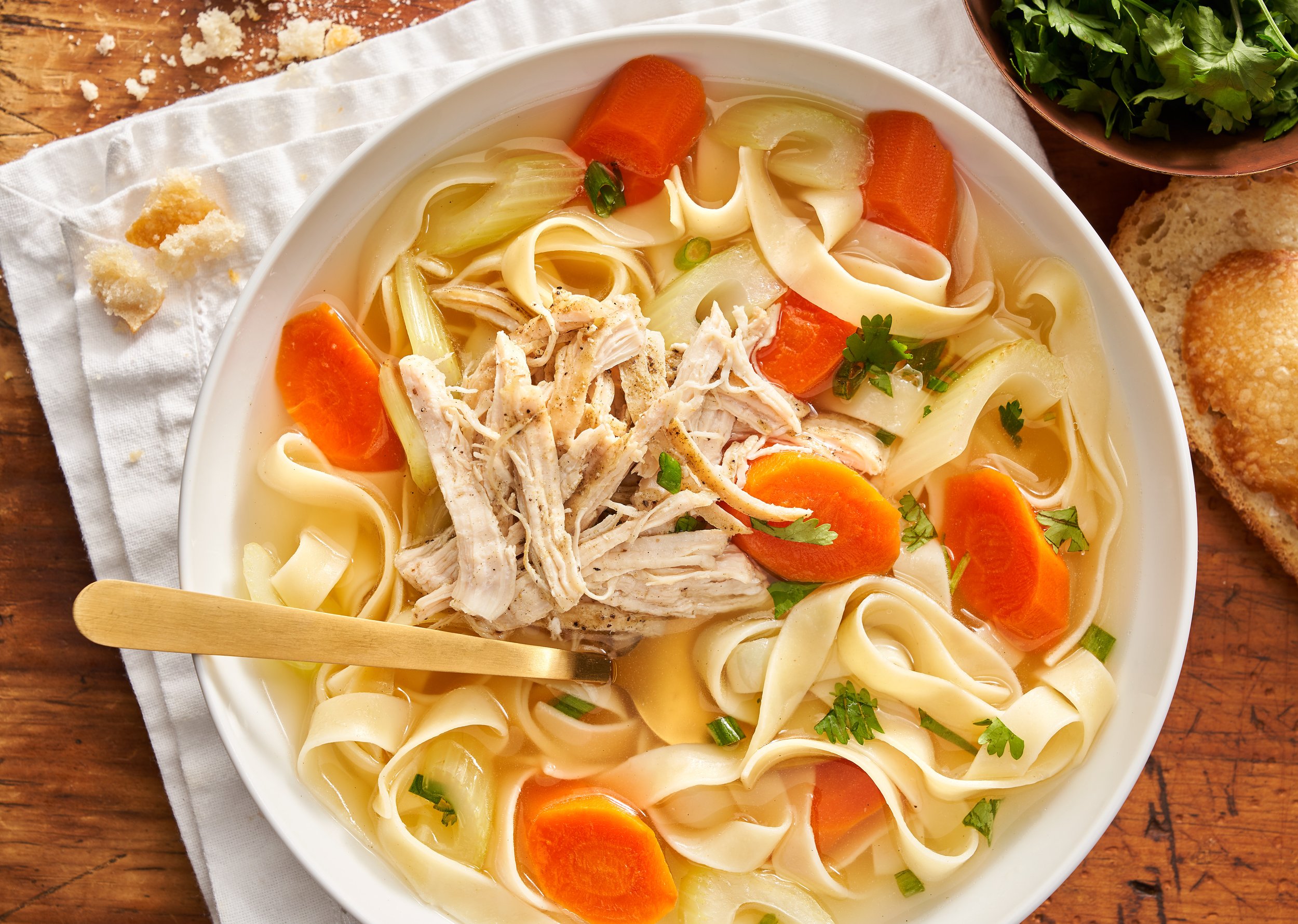 Bowl of chicken noodle soup with carrots, celery, shredded chicken, and herbs on a wooden table.