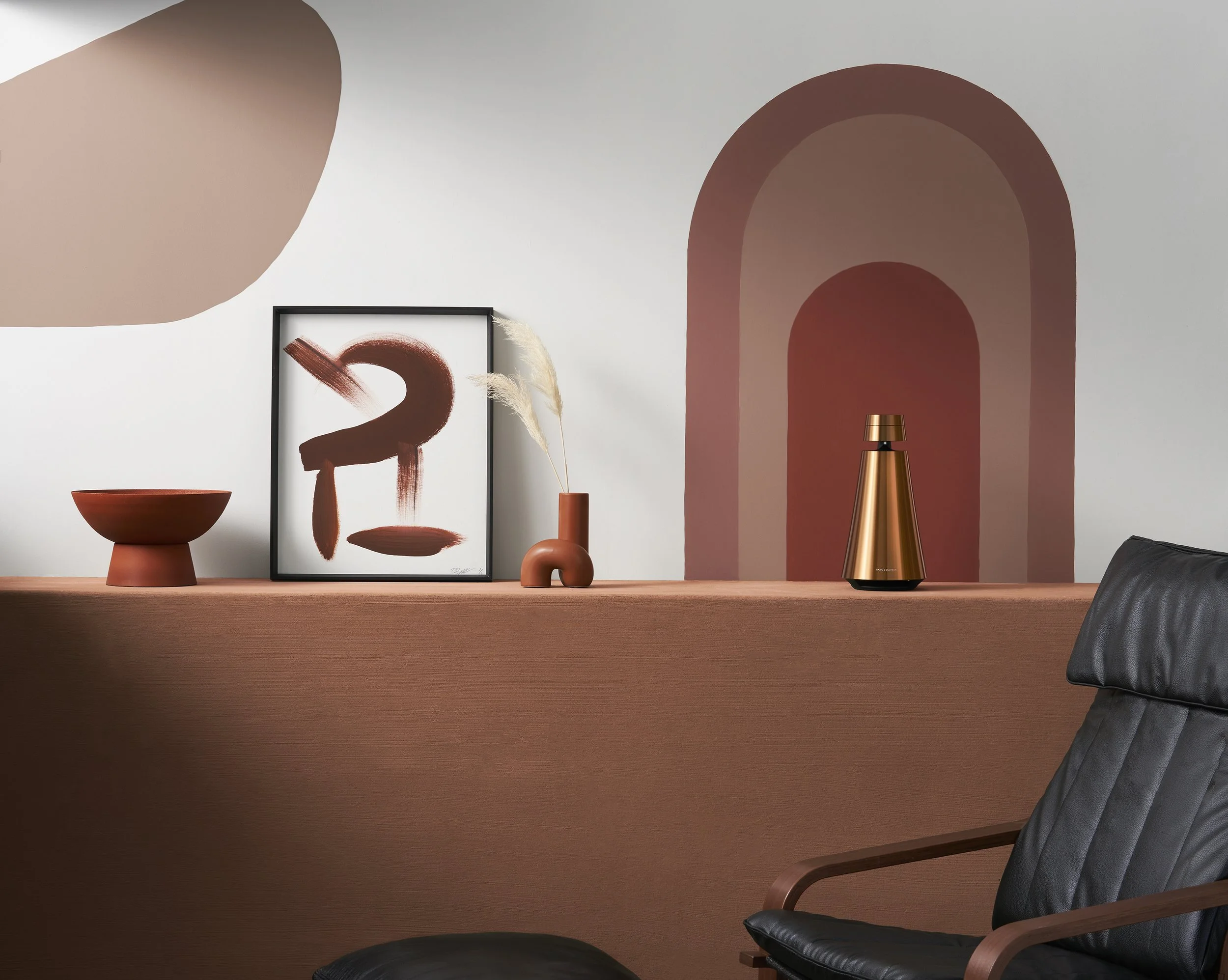 A minimalist room with a wooden desk, modern black leather chair, and decorative items including framed art, vases, and a copper table lamp. The decor features earthy tones and accent colors like rust and beige, with a large arched wall design in the