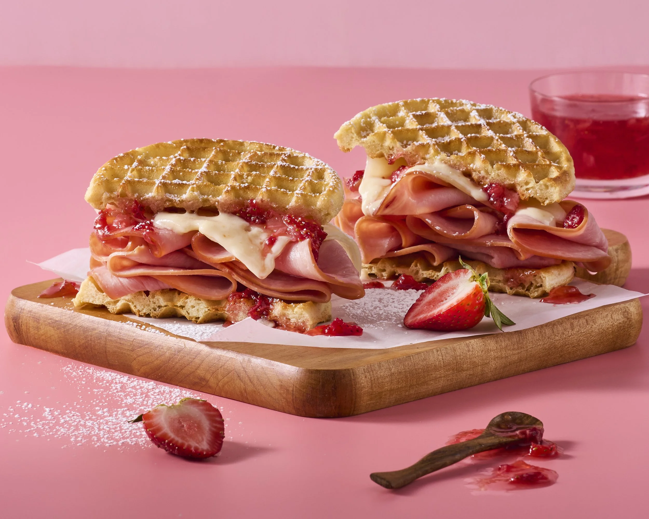 Waffles filled with ham, cheese, and strawberry jam on a wooden board with strawberries and a glass of jam in the background.