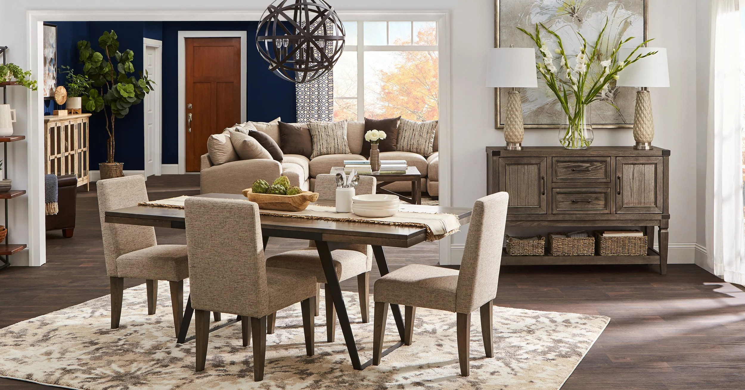 A dining area with a wooden table and four beige upholstered chairs on a patterned rug, overlooking a living room with a beige sectional sofa, pillows, a coffee table, and a large window with autumn trees outside. There's a wooden sideboard with lamp