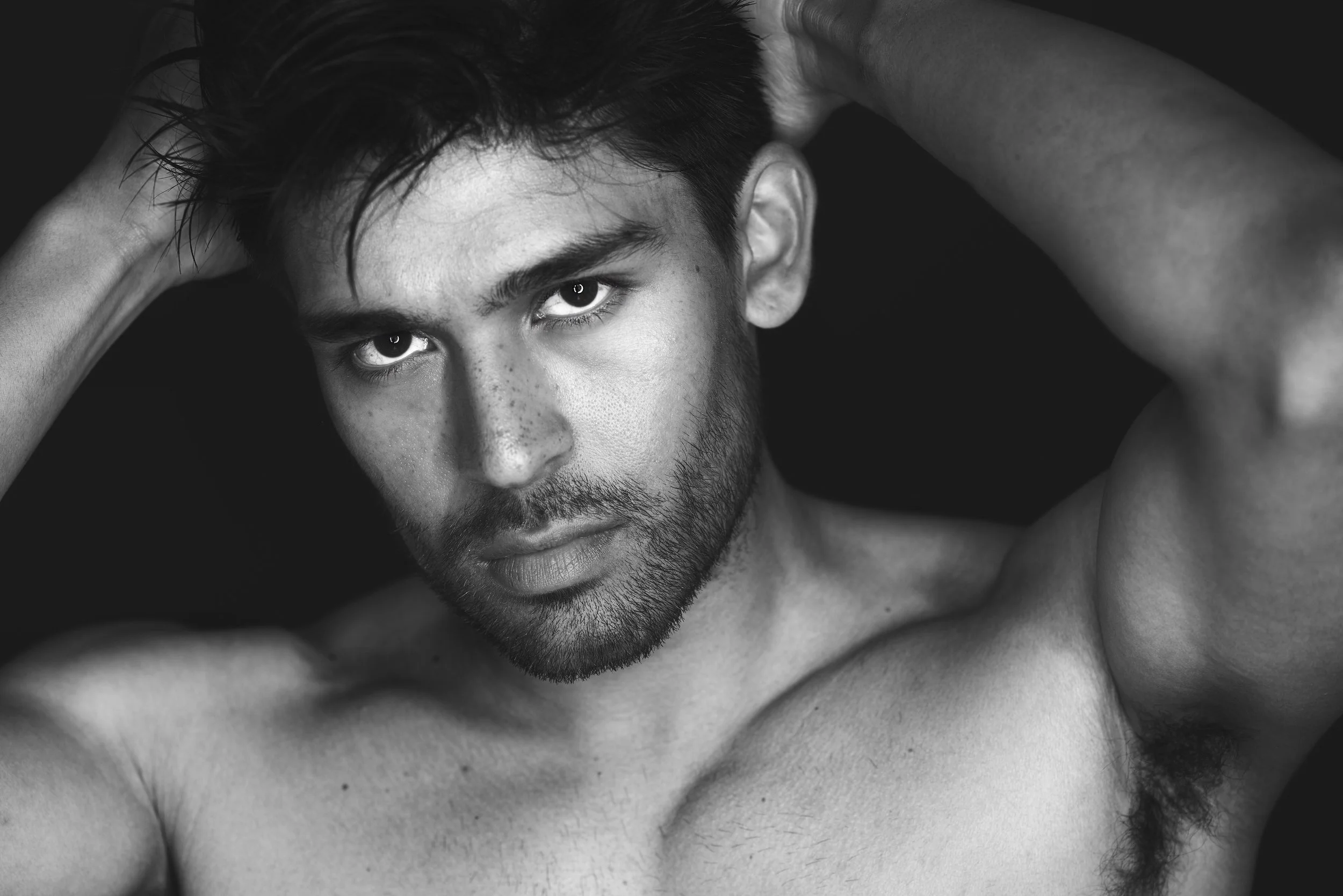 A black and white portrait of a shirtless man with dark hair, a beard, and intense eyes, looking directly at the camera with hands behind his head.