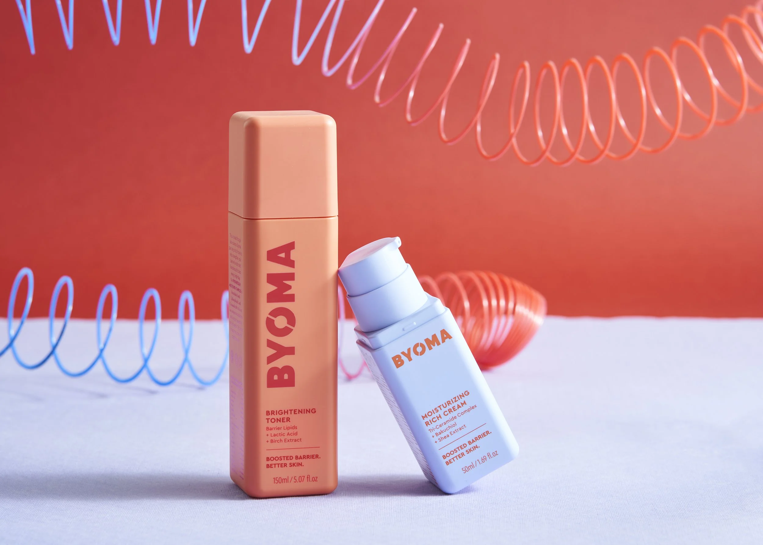 Two skincare products from BYOMA, a brightening toner and a moisturizing rich cream, are displayed on a white surface with a colorful abstract background of coiled spirals in pink and blue.
