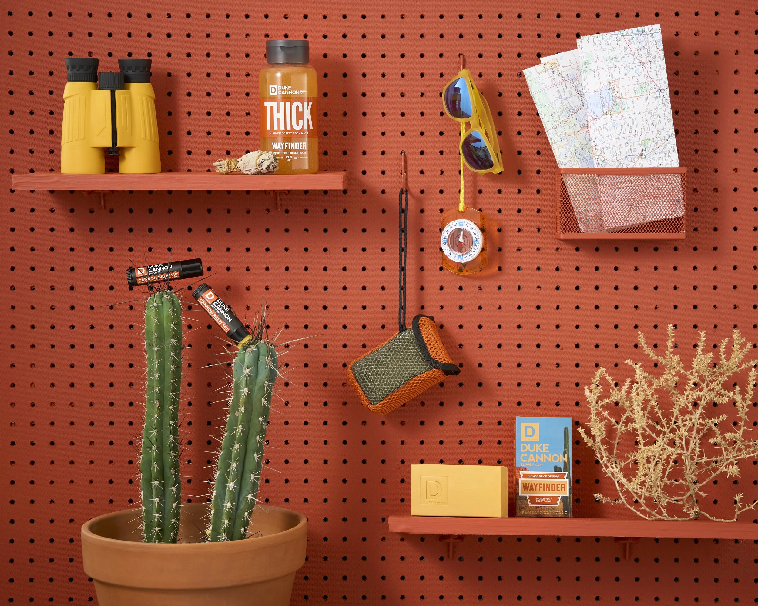 Organized camping gear on a red pegboard wall, including a yellow binocular case, a bottle of Duke Cannon Thick Body Wash, sunglasses, a map, a compass, a small pouch, a yellow box, a brochure, and a potted cactus and succulent.