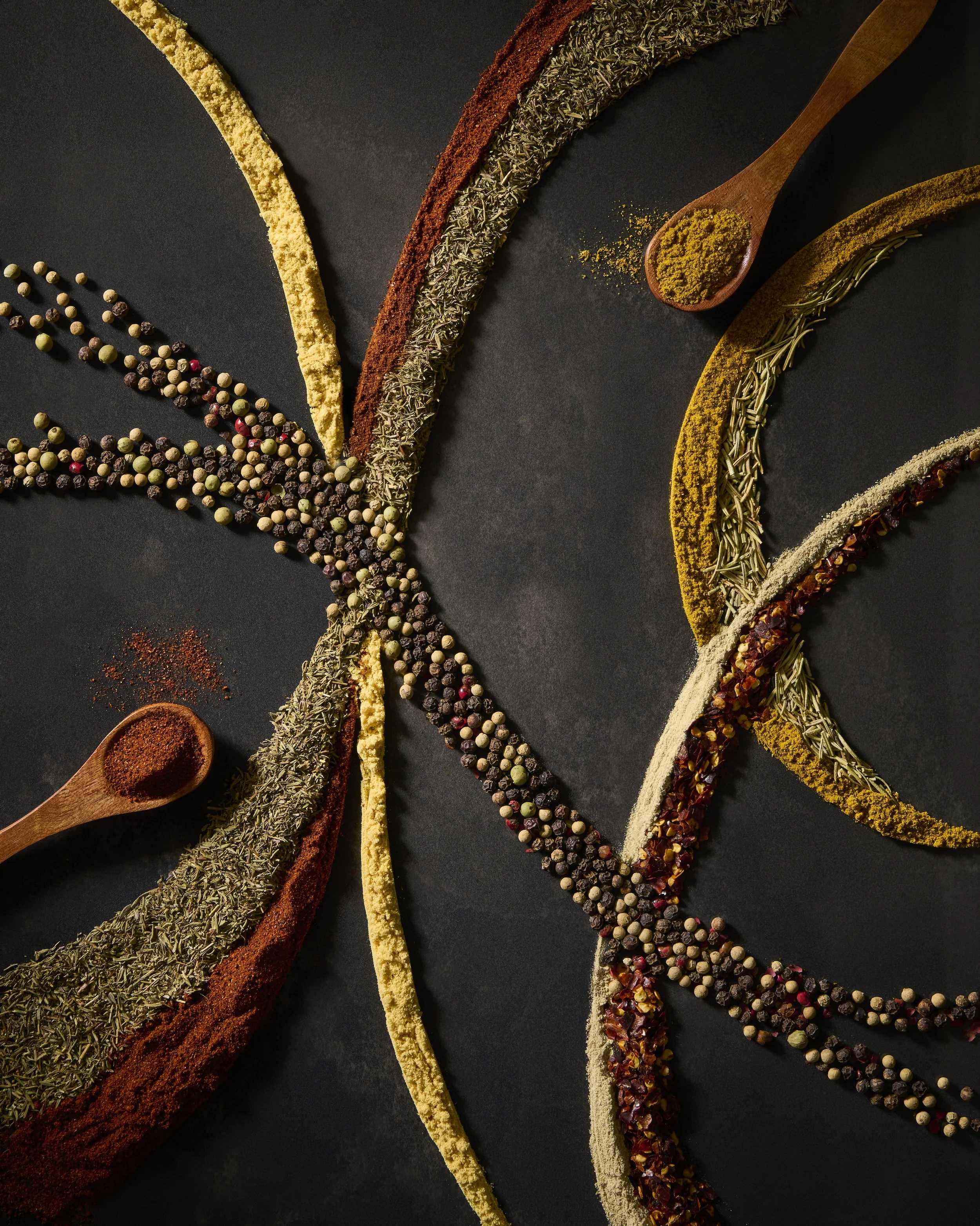 Assorted spices and herbs arranged in curved lines on a black background, with small wooden spoons holding ground spices.