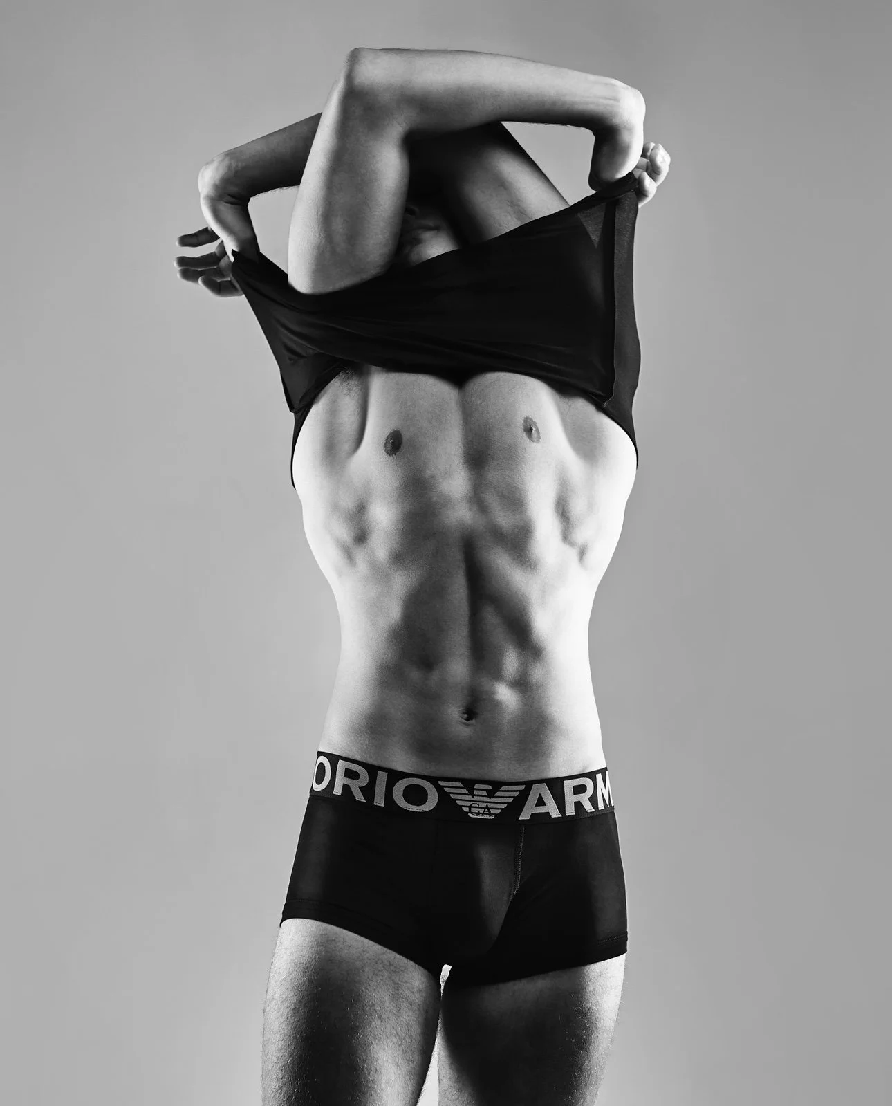 A man lifting his shirt to reveal a muscular abdomen while wearing Armani underwear, against a plain background.