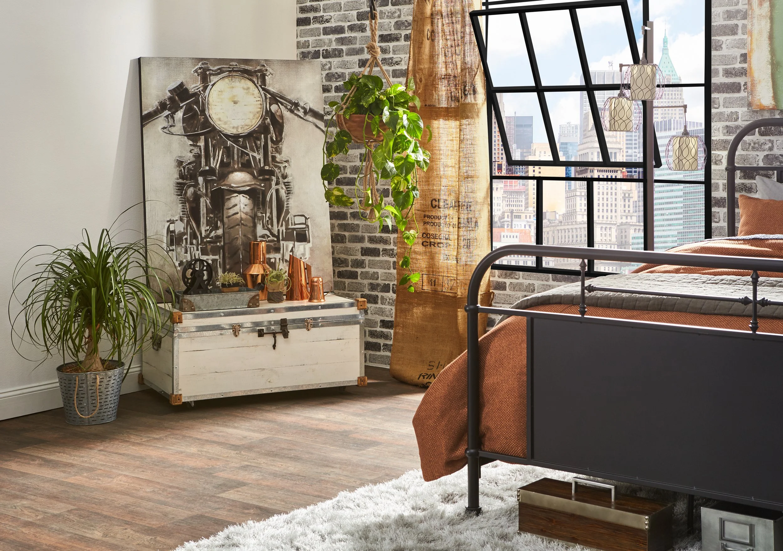 Decorated bedroom with large window overlooking cityscape, black metal bed frame with rust-colored bedding, plants, artwork, and rustic decor.
