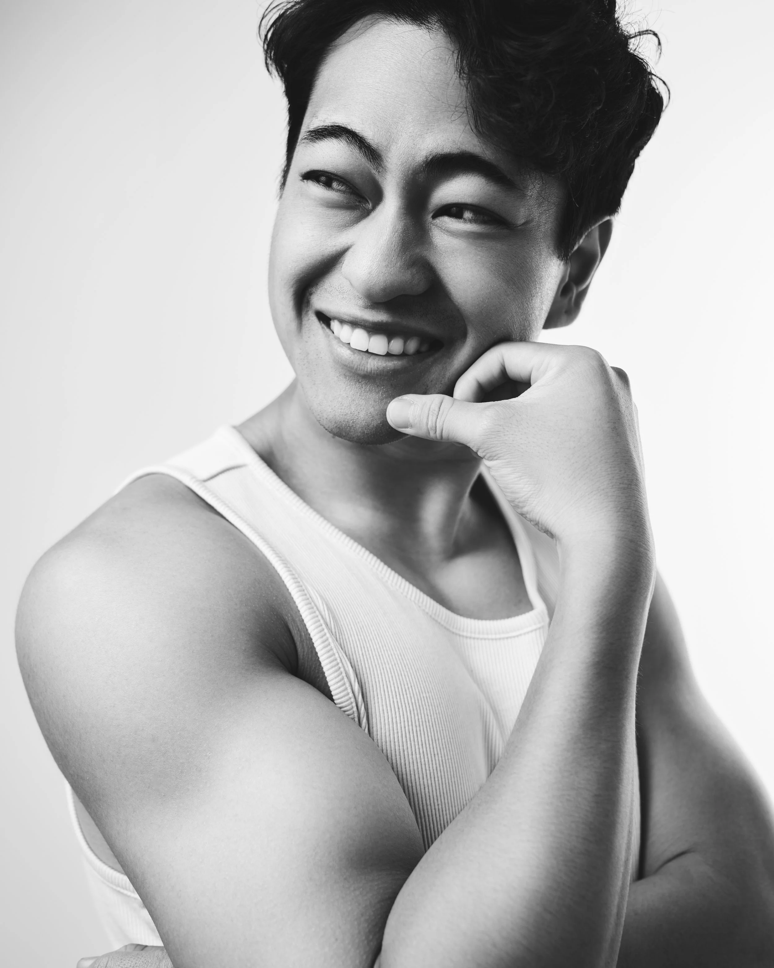 Black and white photo of a smiling man with short dark hair, wearing a sleeveless shirt, resting his chin on his hand, showing his bicep.