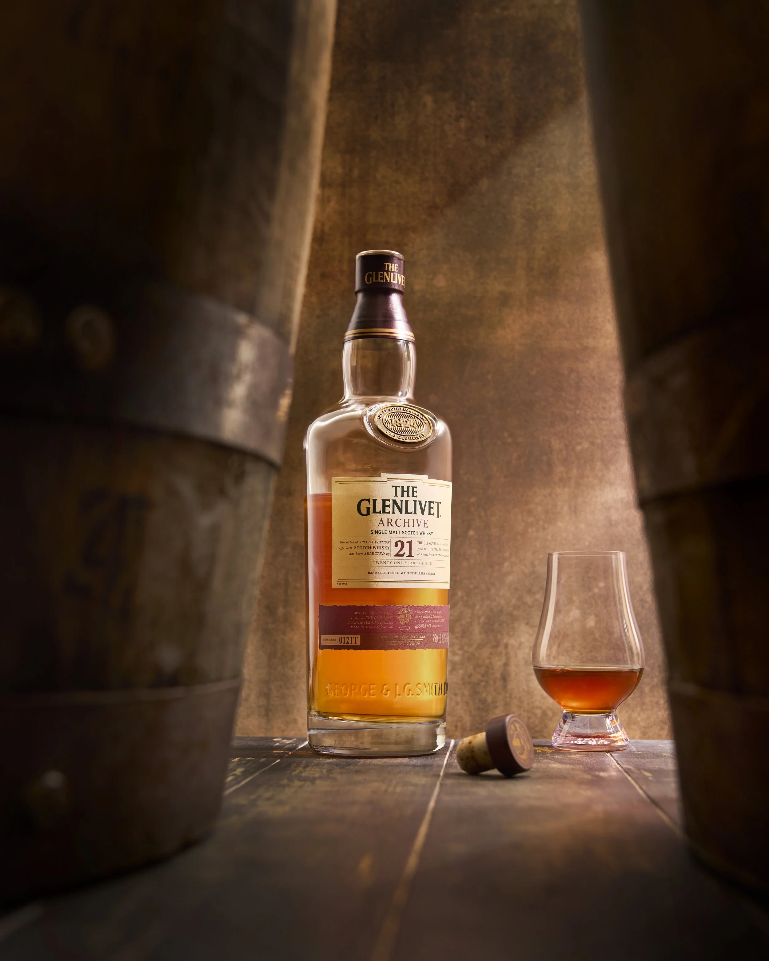 A bottle of The Glenlivet Archive 21-year-old single malt Scotch whisky, a whisky glass filled with whisky, and a cork lie on a wooden surface, framed by barrels in a dimly lit setting.