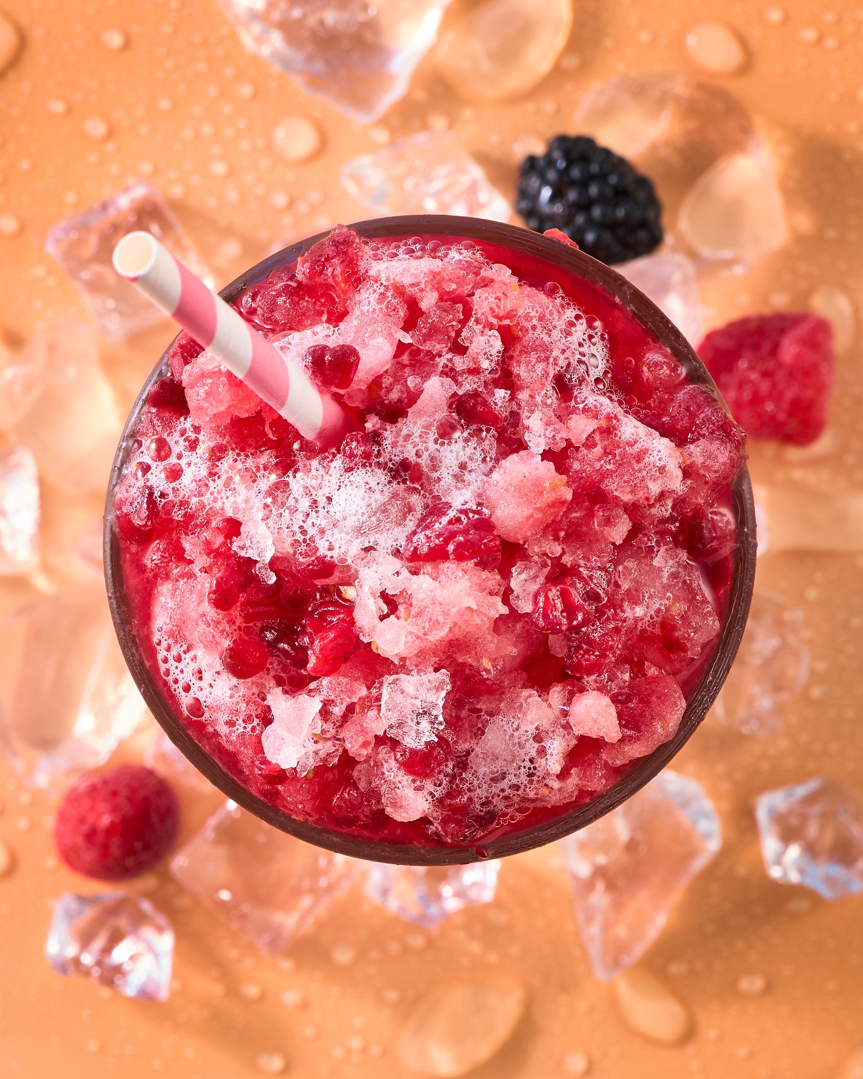 Close-up of a red flavored frozen beverage with crushed ice and a striped straw, on a background of ice cubes and blackberries.
