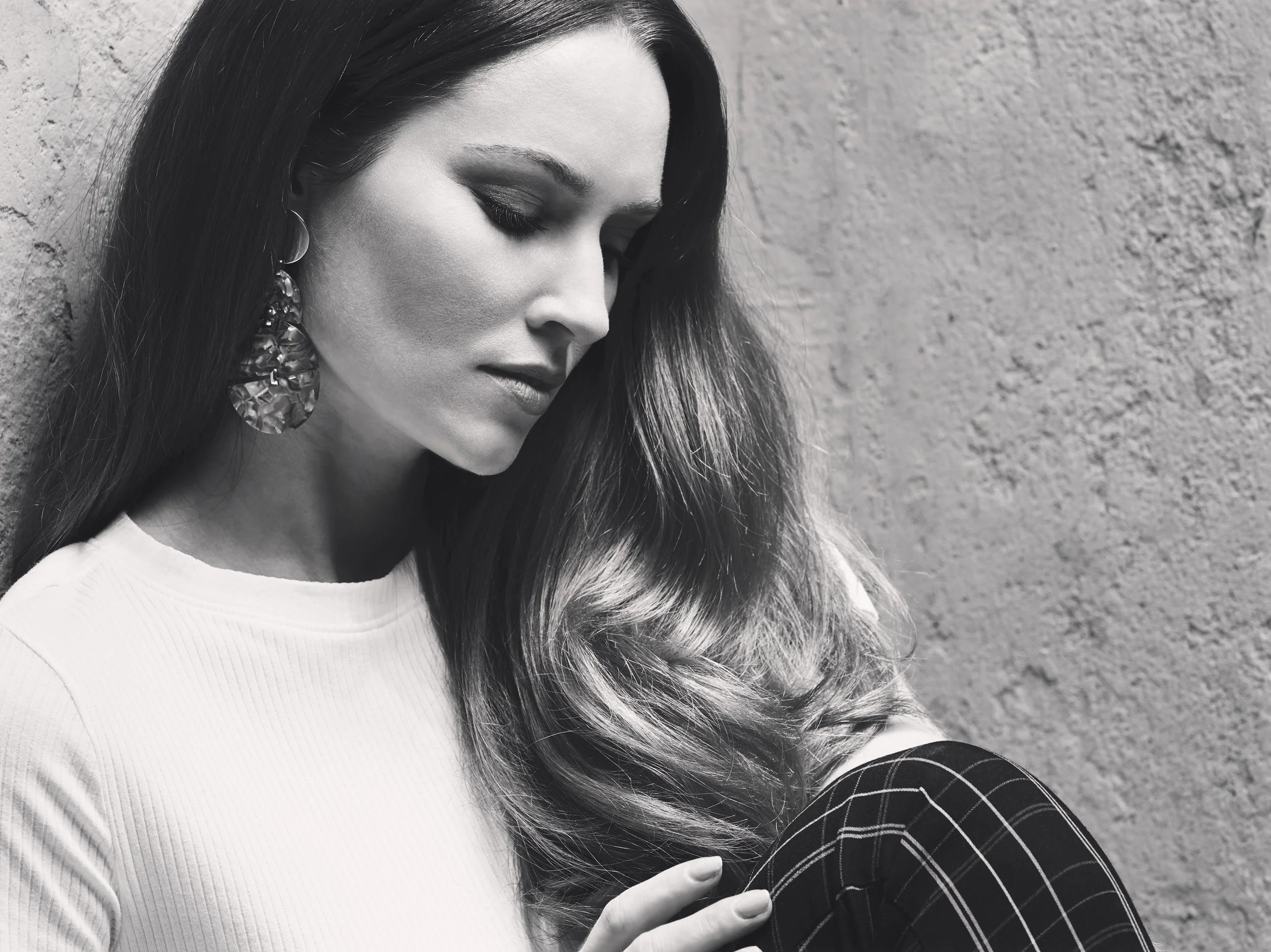 A woman with long hair, wearing earrings and a white top, is looking down while sitting against a textured wall.