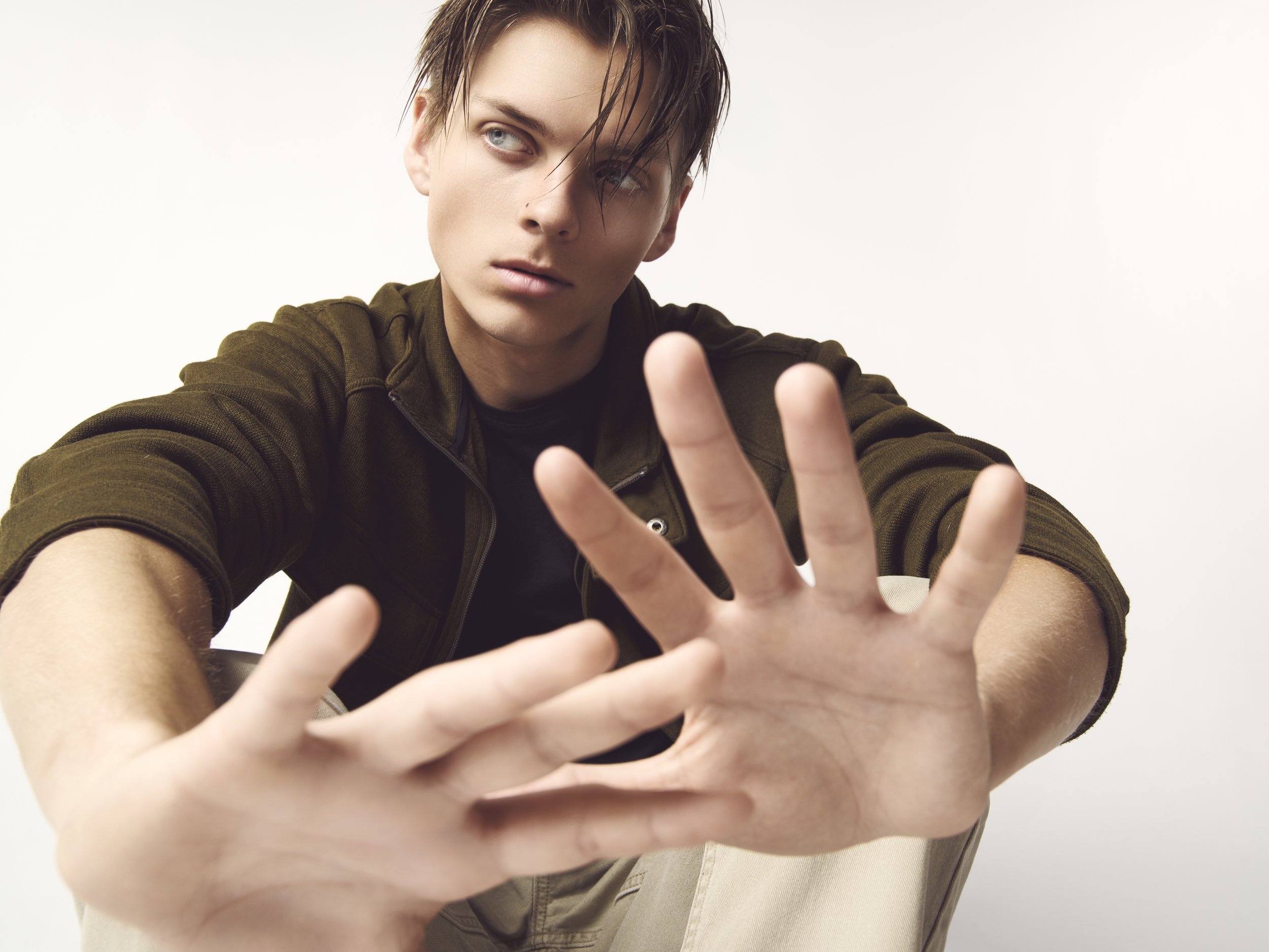 A young man with short dark hair and light skin extends his hands towards the camera, with fingers spread apart, as he looks slightly to the side with a neutral expression. He wears a dark green jacket and a black shirt, sitting against a plain white