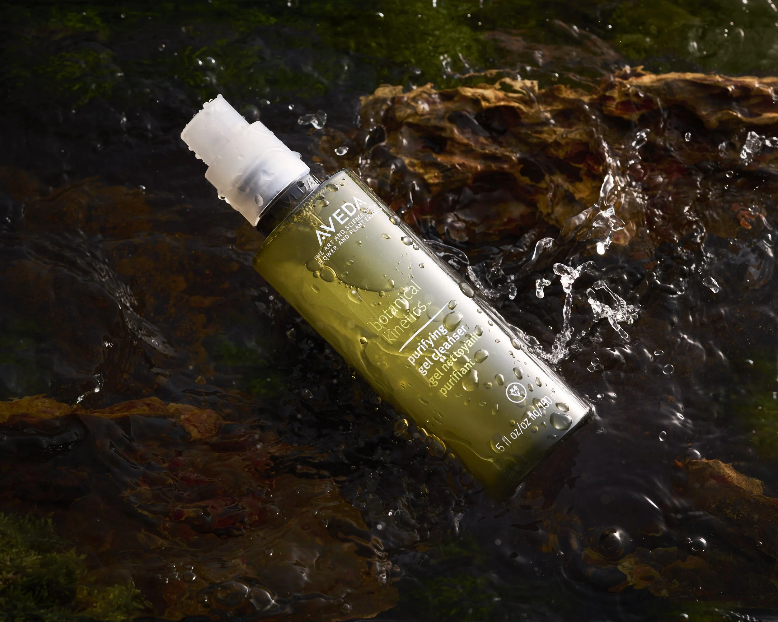 A green bottle of Aveda botanical kinetic purifying gel cleanser lying in a stream with water flowing around it and mossy rocks.