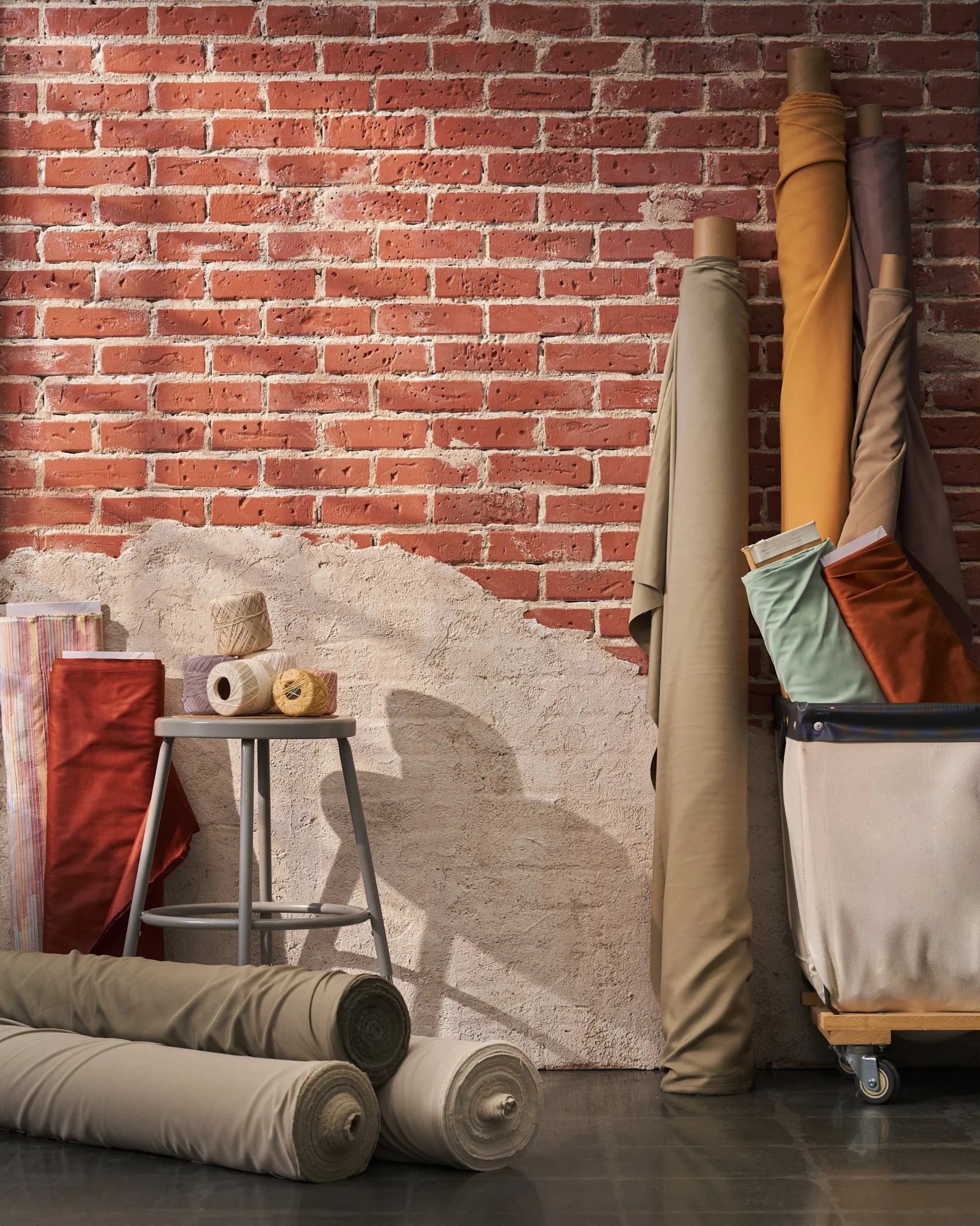 A variety of fabric rolls, yarns, and textiles in front of a brick and textured wall, with fabric rolls leaning against a cart.