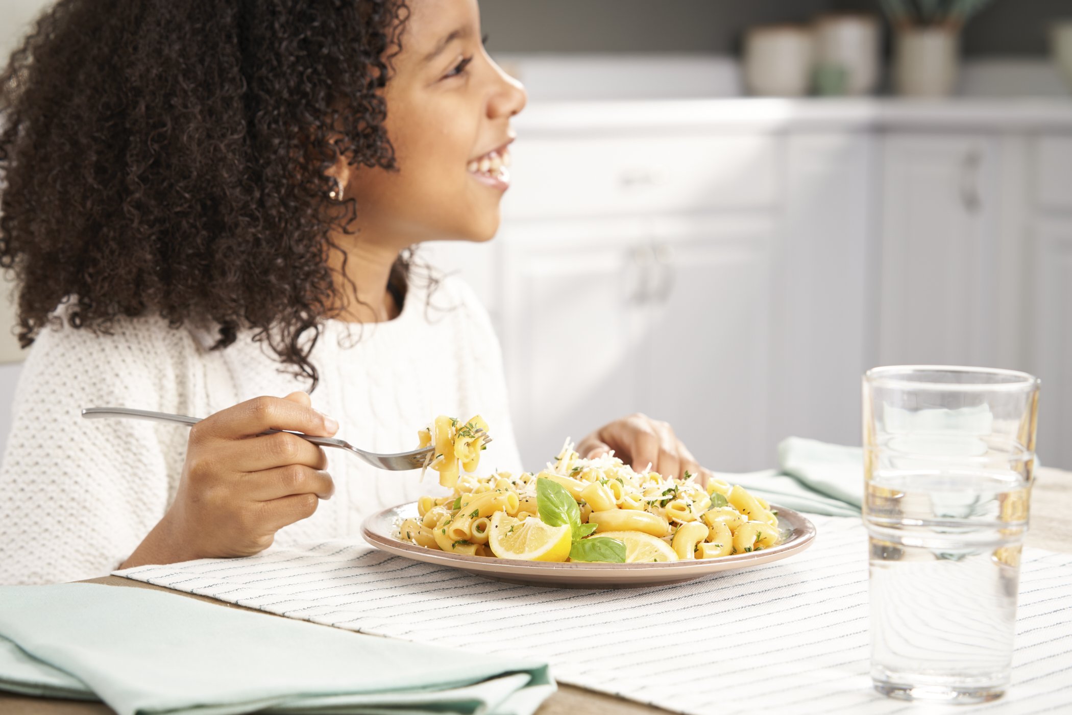 A young woman with curly hair enjoying a plate of pasta with lemon slices and basil, sitting at a dining table with a glass of water nearby.