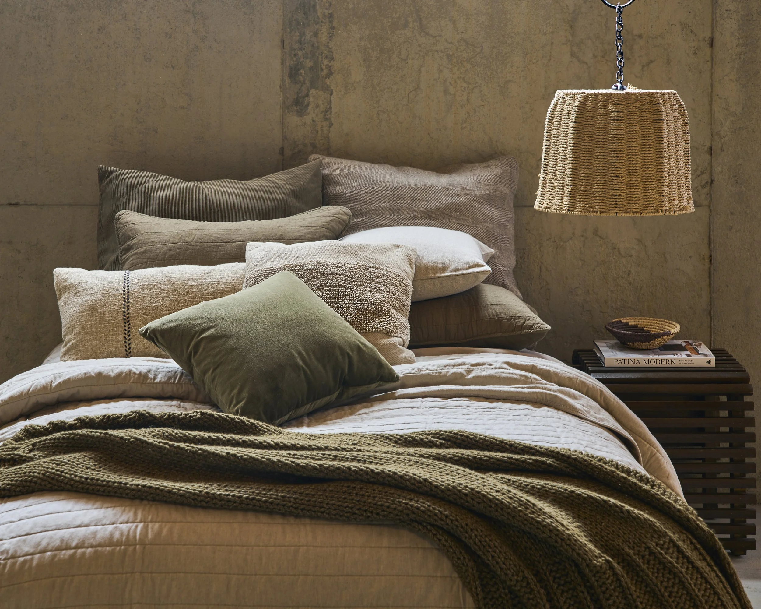 A cozy bed with a variety of neutral-colored pillows and a chunky knit blanket, beside a dark wooden slatted nightstand holding magazines and a small woven basket; hanging wicker light fixture above.