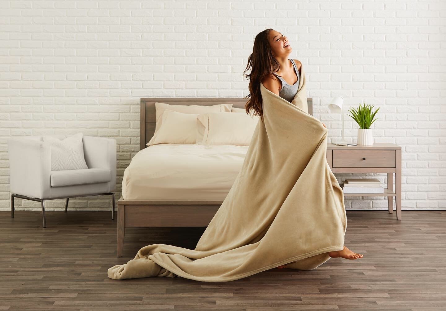 A woman wrapped in a beige blanket standing in a bedroom, smiling and looking away, with a bed, a white armchair, a bedside table with a plant and a lamp, and a white brick wall in the background.