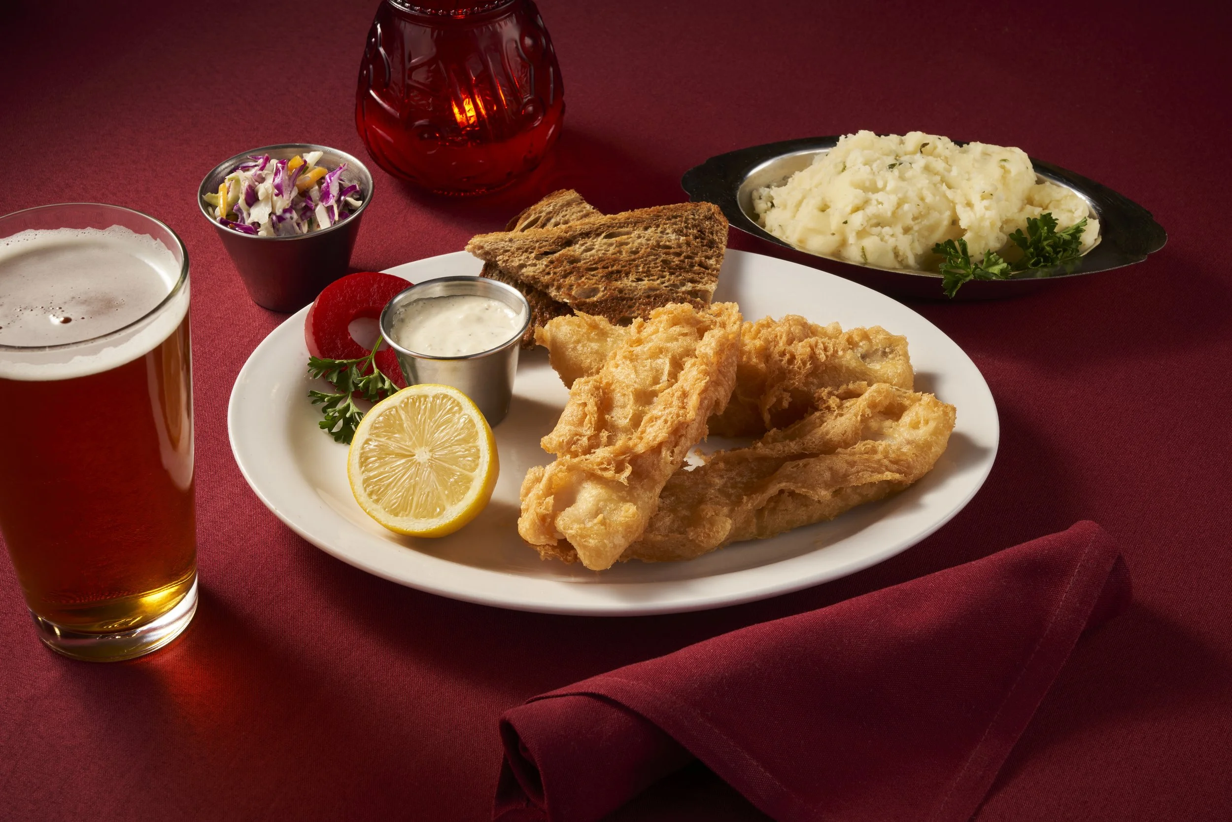 Fried fish with lemon, coleslaw, mashed potatoes, toast, and a glass of beer on a red tablecloth.