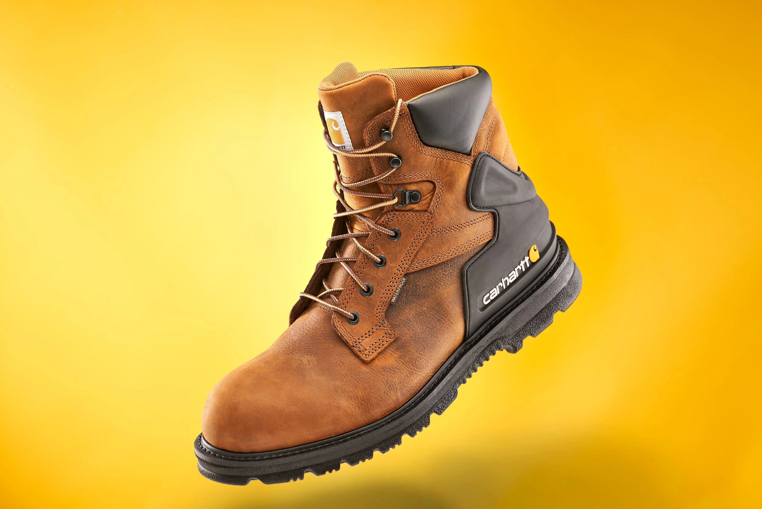 A brown Carhartt work boot with black rubber heel and sole, displayed against a yellow background.