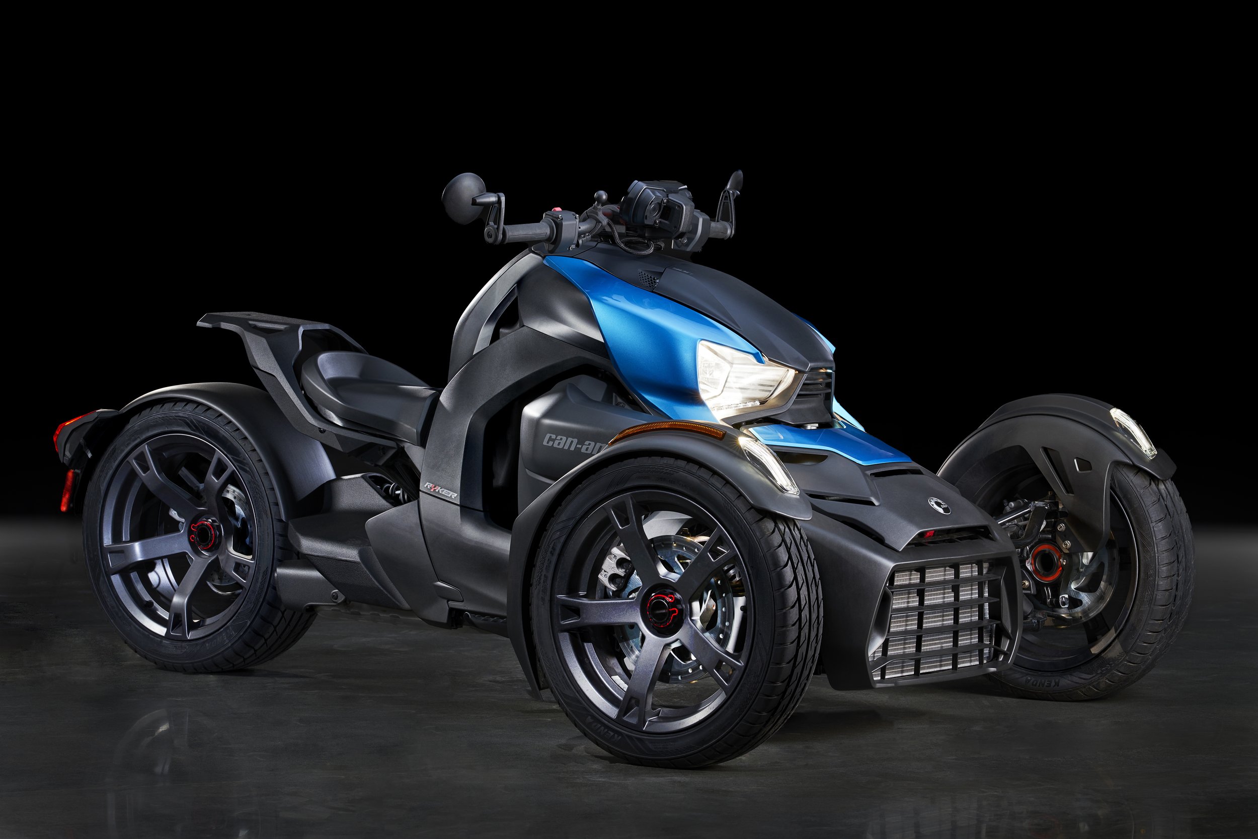 A three-wheeled Can-Am off-road vehicle with one large front wheel and two smaller rear wheels, featuring a blue and black color scheme, black tires, and a single seat on a dark background.