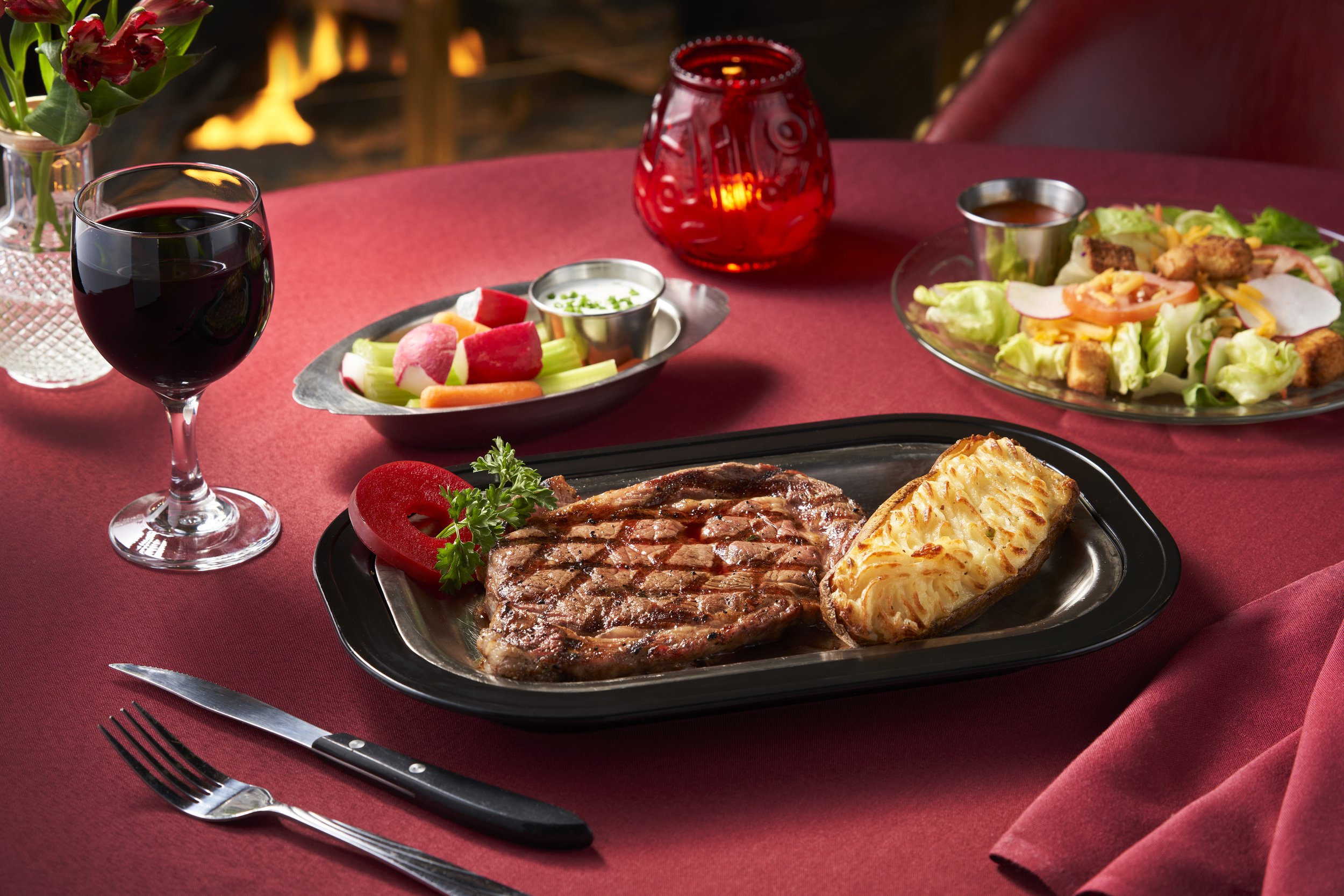 A dinner plate with grilled steak, mashed potatoes, and a red bell pepper garnish, set on a red tablecloth. In the background, there is a bowl of fresh vegetables, a salad with lettuce, tomatoes, and croutons, a glass of red wine, and a candle holder