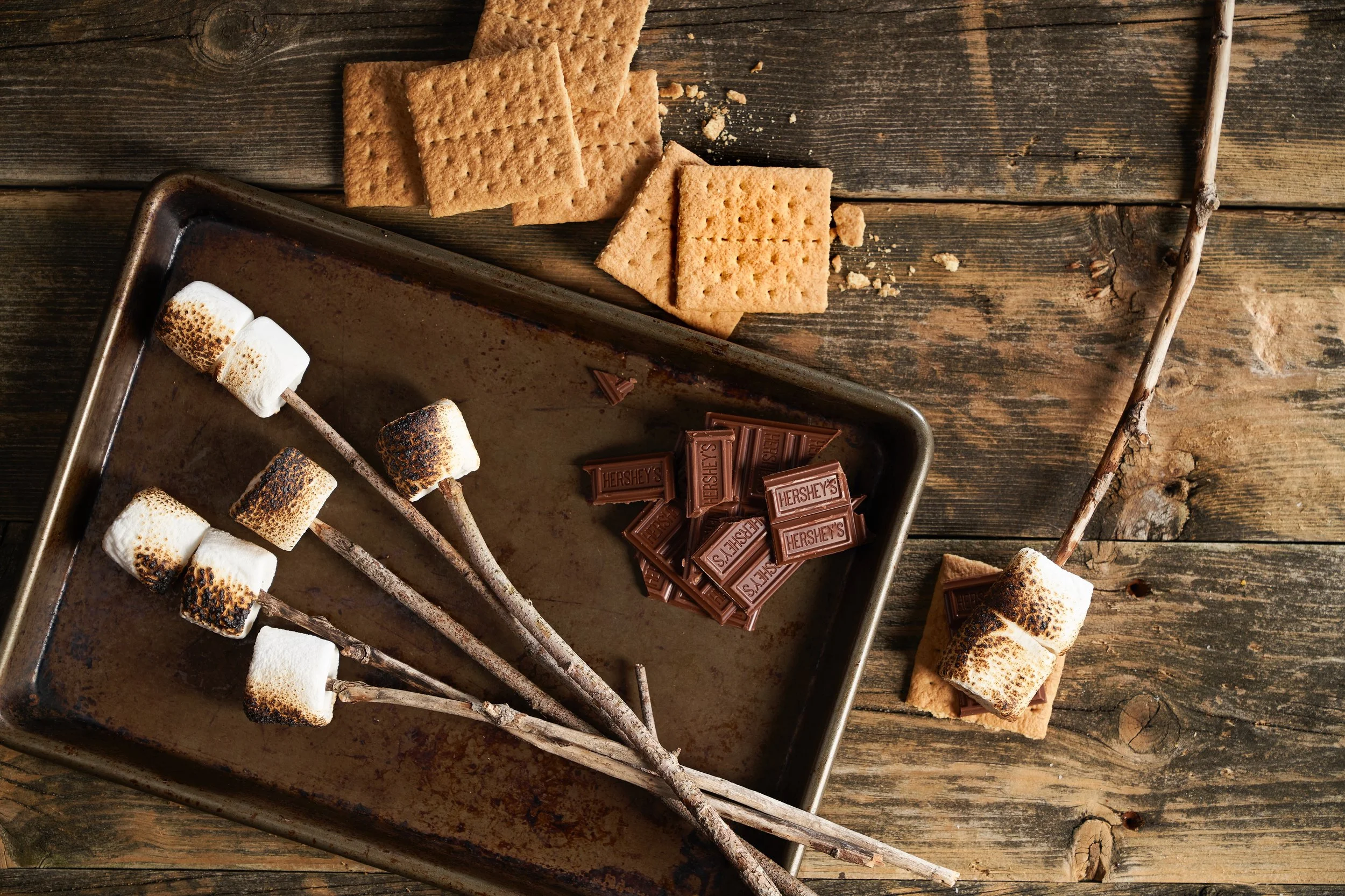 S'mores ingredients on a rustic wooden table: roasted marshmallows on sticks, chocolate pieces, graham crackers, with some graham crackers and broken graham crackers nearby.