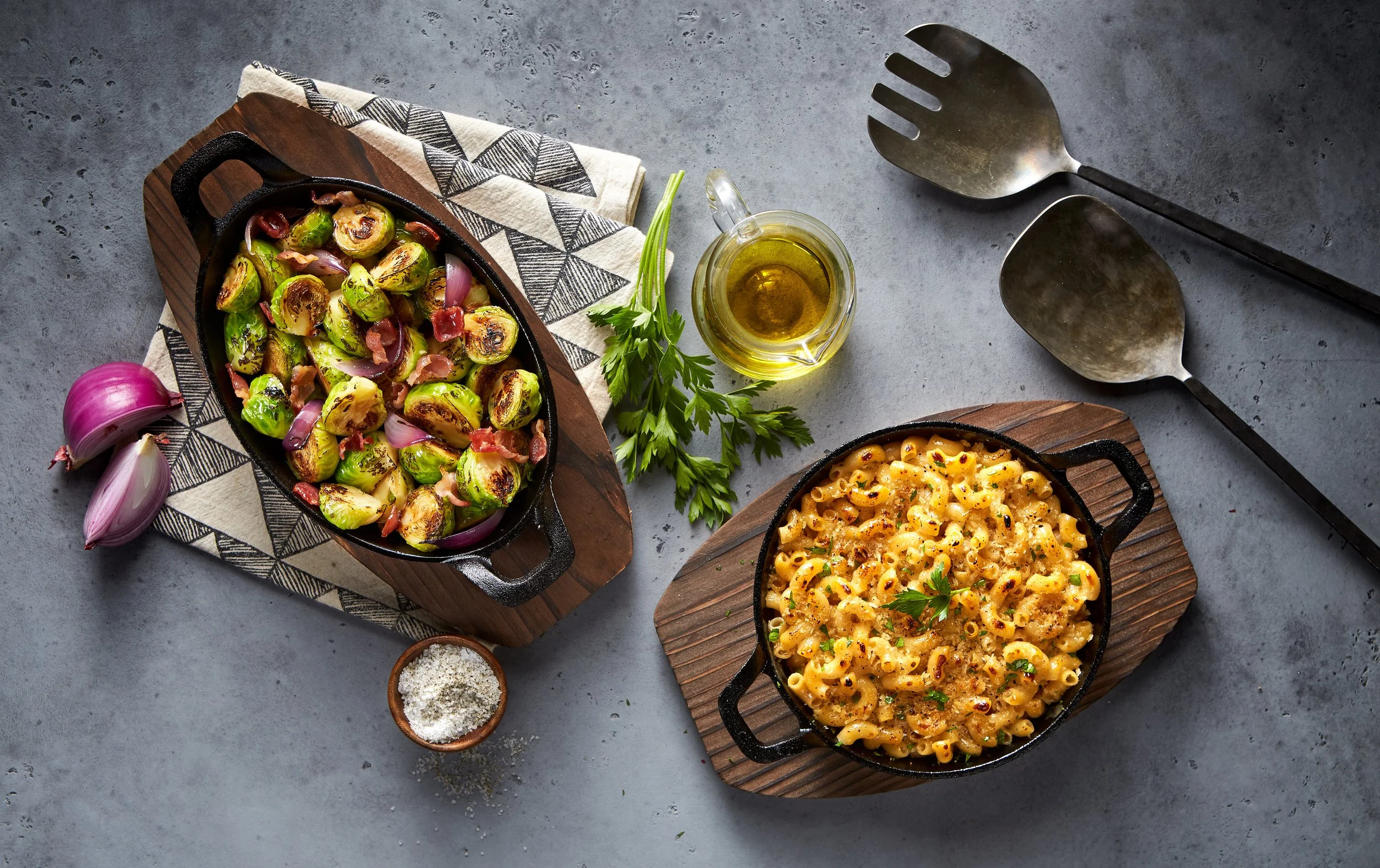 Cast iron dishes with roasted Brussels sprouts and macaroni and cheese, garnished with parsley, with a bowl of salt, a bottle of olive oil, and fresh parsley on a gray surface.