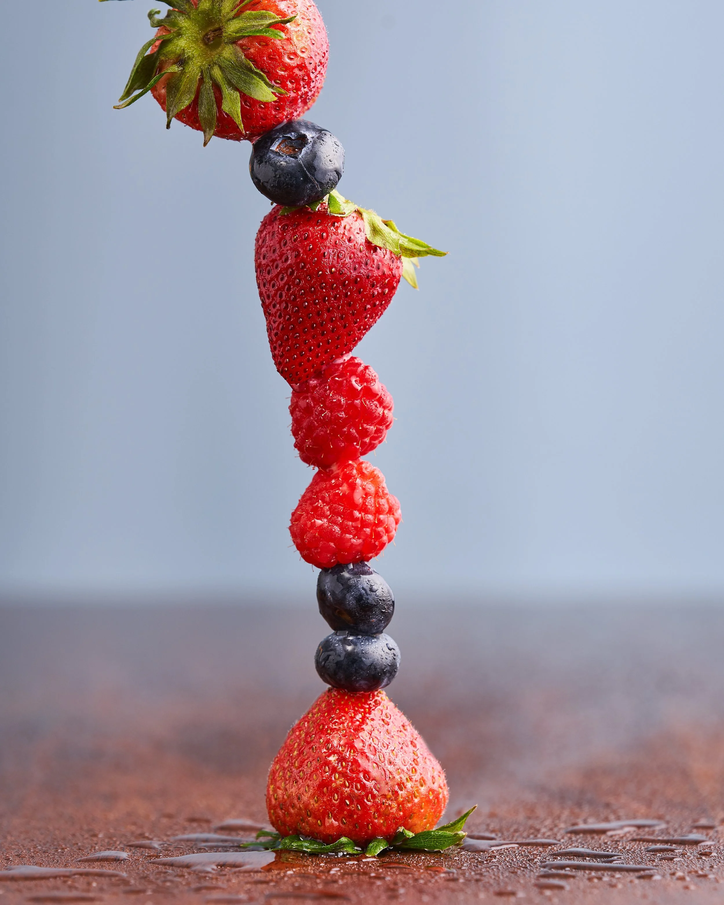 A vertical arrangement of various fresh berries, including strawberries, blueberries, raspberries, and blackberries, balanced on a wet surface.