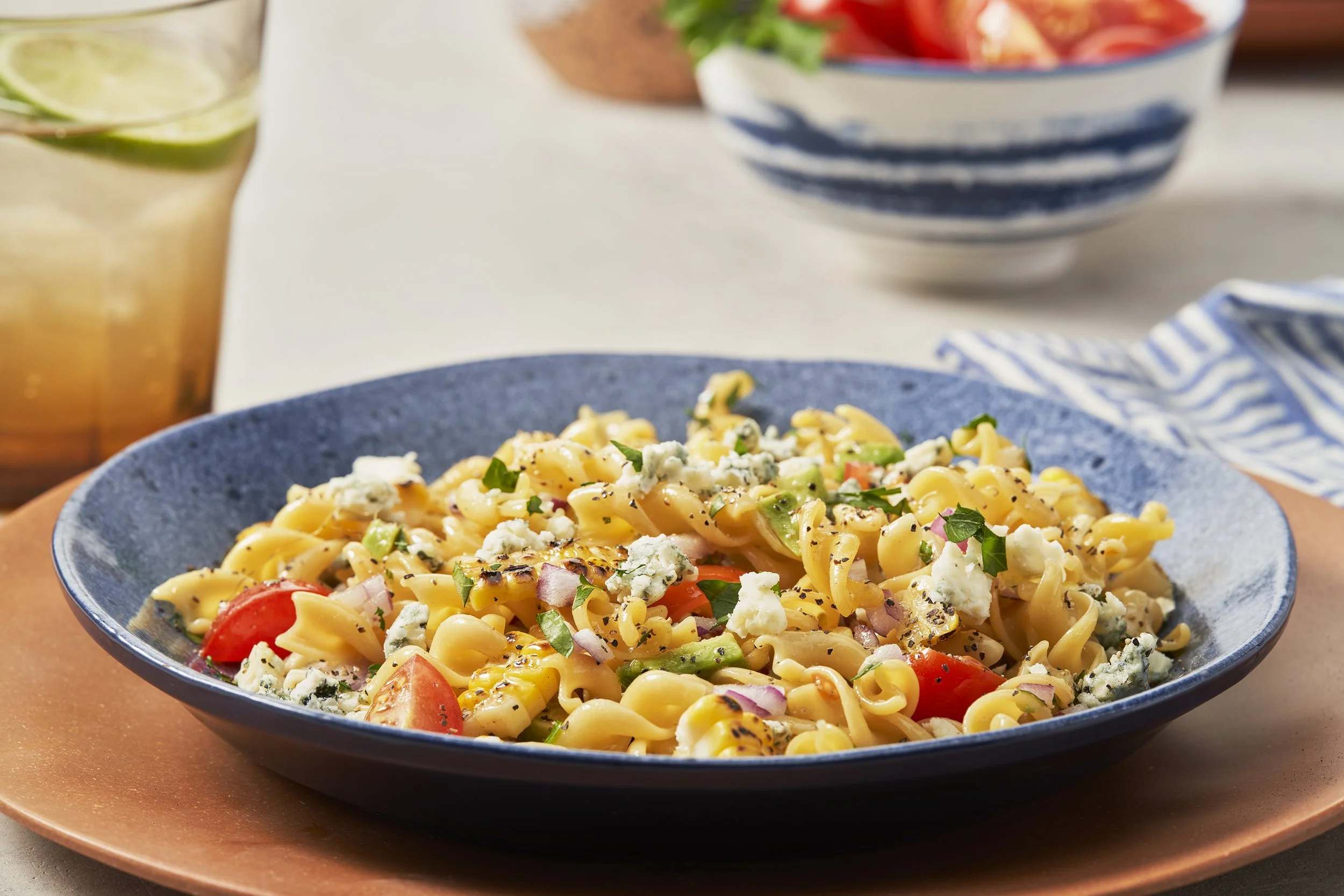 Close-up of a blue bowl filled with pasta salad containing rotini pasta, cherry tomatoes, chopped red onions, and crumbled blue cheese, garnished with chopped herbs. A glass with lime slices and a cold beverage is in the background. There is also a b