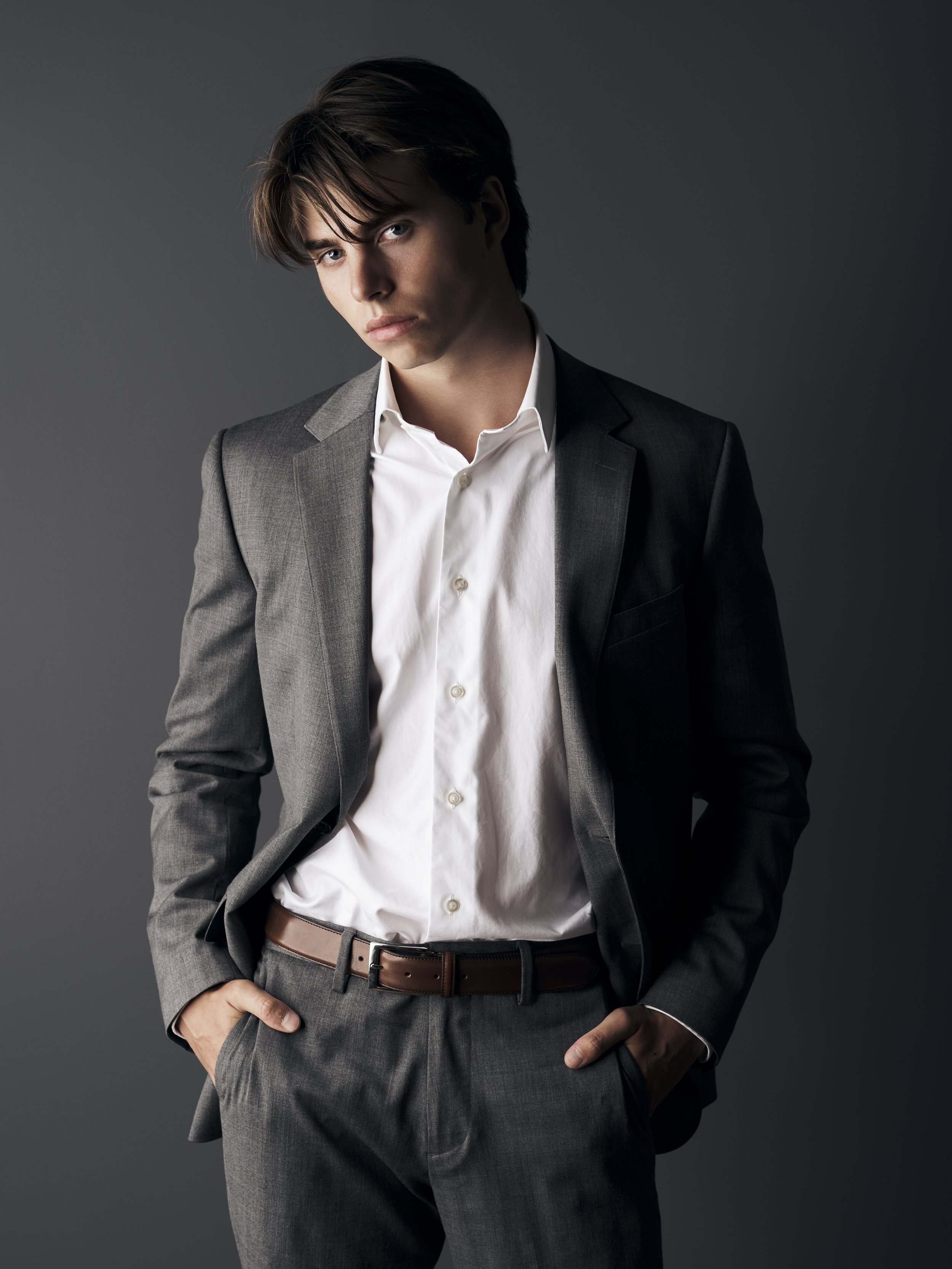 Young man with dark hair wearing a gray suit and white shirt, standing against a dark background.