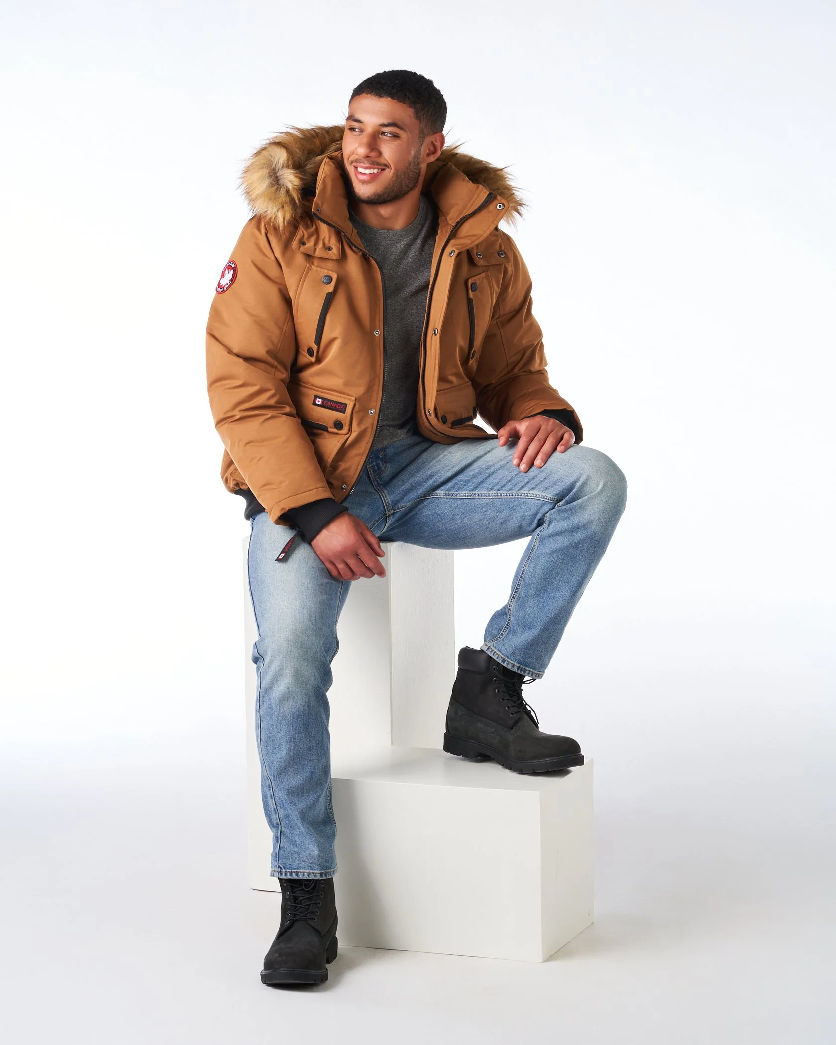 A young man with dark hair and beard sitting on a white platform and smiling, wearing a brown winter coat with a hood and fur lining, blue jeans, and black boots, against a white background.