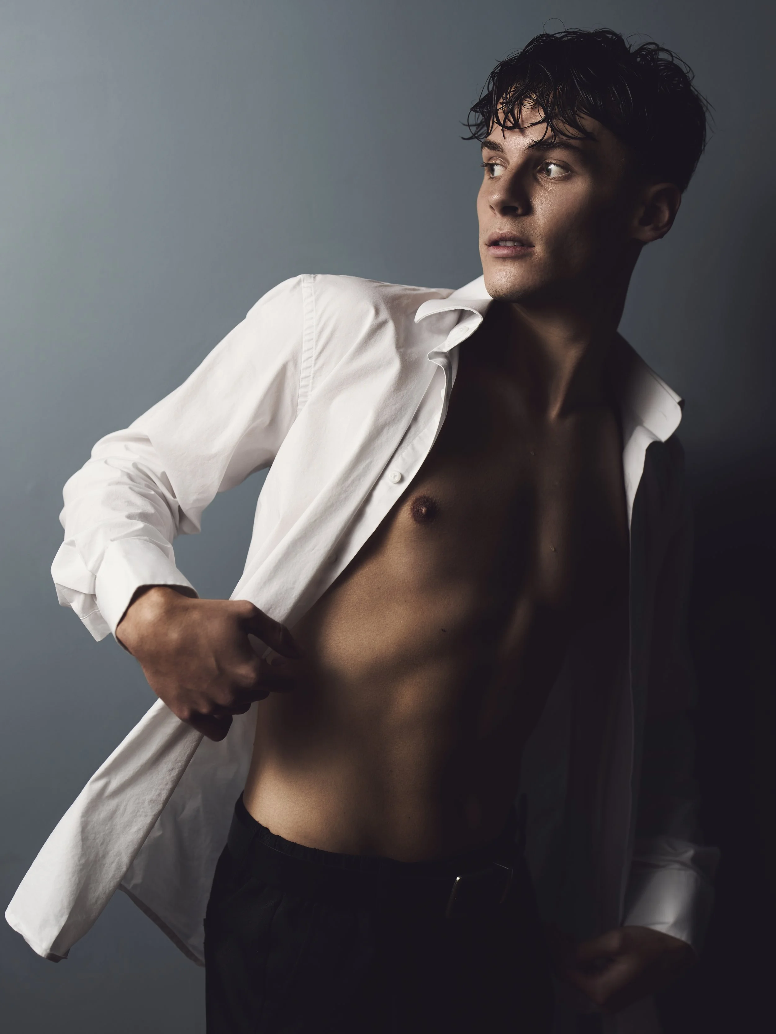 A young man with dark, wet-looking hair is partially undressed, wearing an open white shirt revealing his bare chest and stomach. He has a serious expression and is posed against a neutral background.