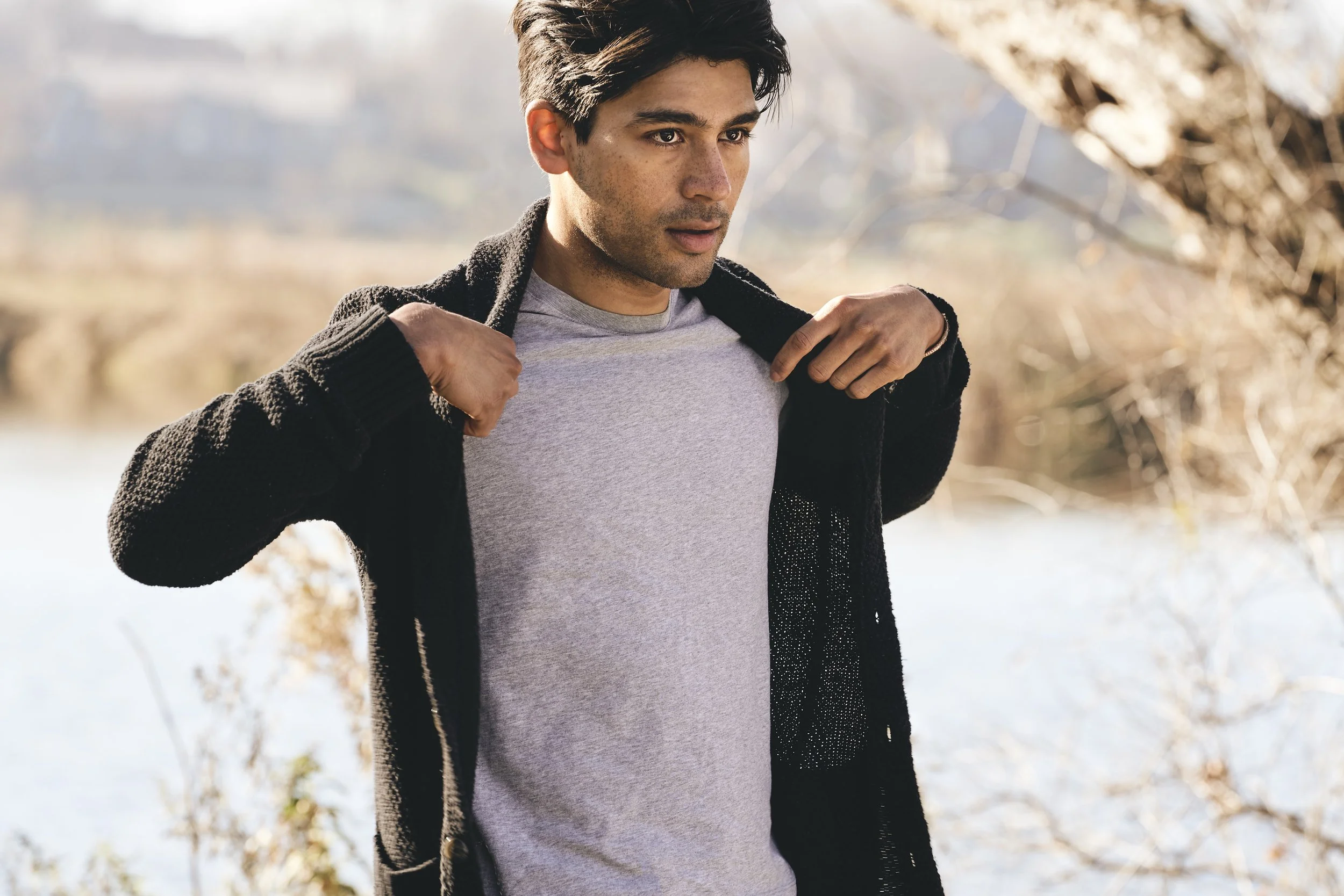 Young man standing outdoors by a lake or river, wearing a gray t-shirt and a black jacket, adjusting his jacket collar, with trees and water in the background.